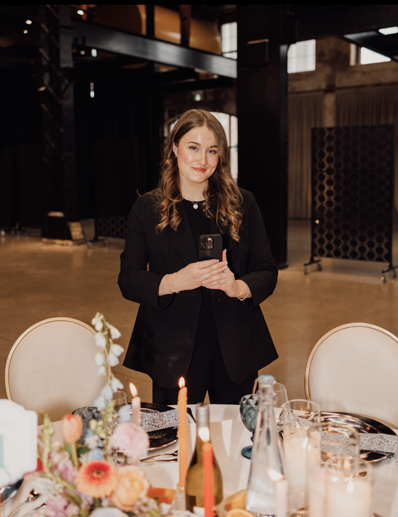 A young woman with long curly hair wearing a black blazer and jewelry, smiling while holding a smartphone at a decorated event table with flowers and candles inside a dimly lit venue.