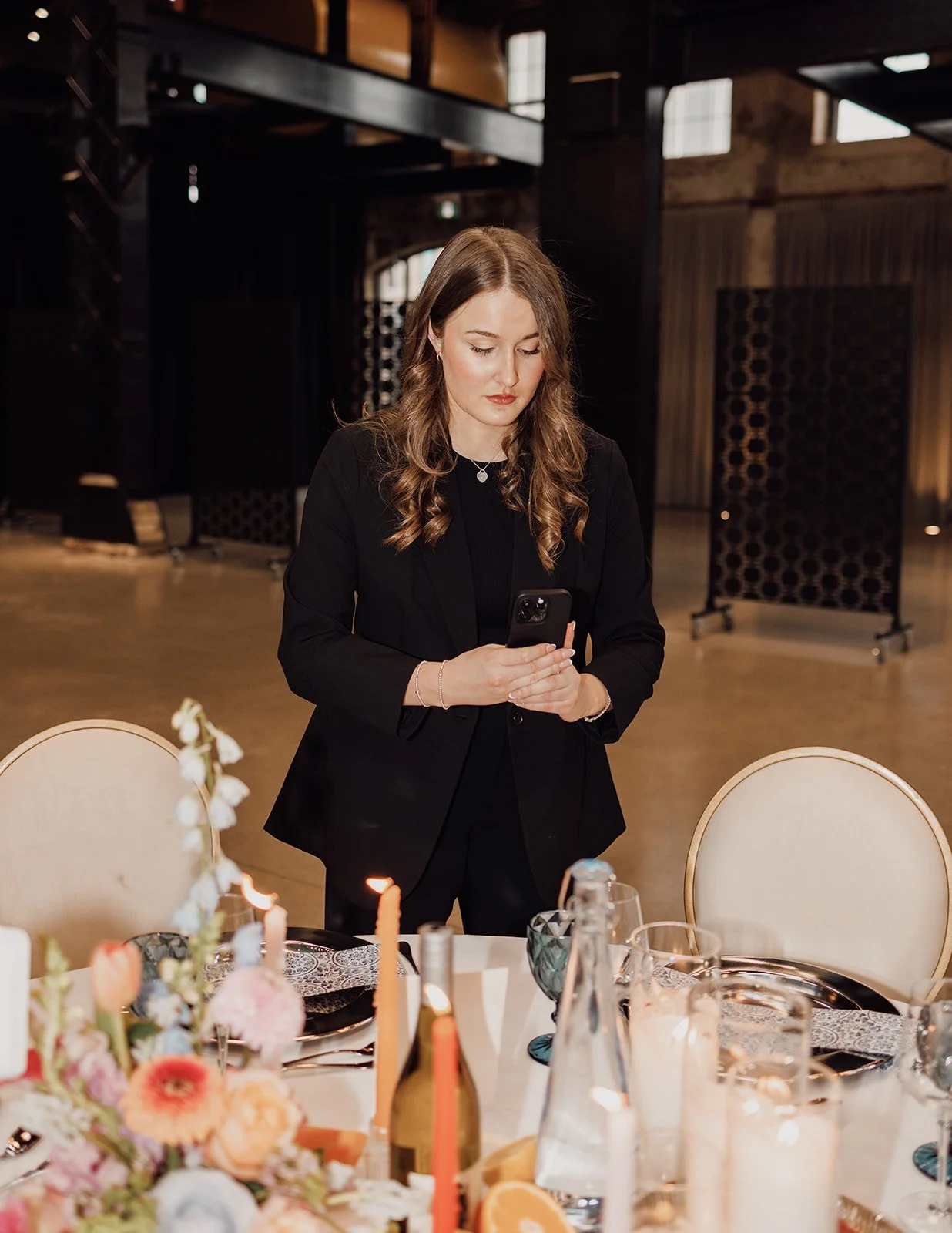 A woman with wavy brown hair in a black blazer standing at a decorated table, looking at her phone in a dimly lit event space.