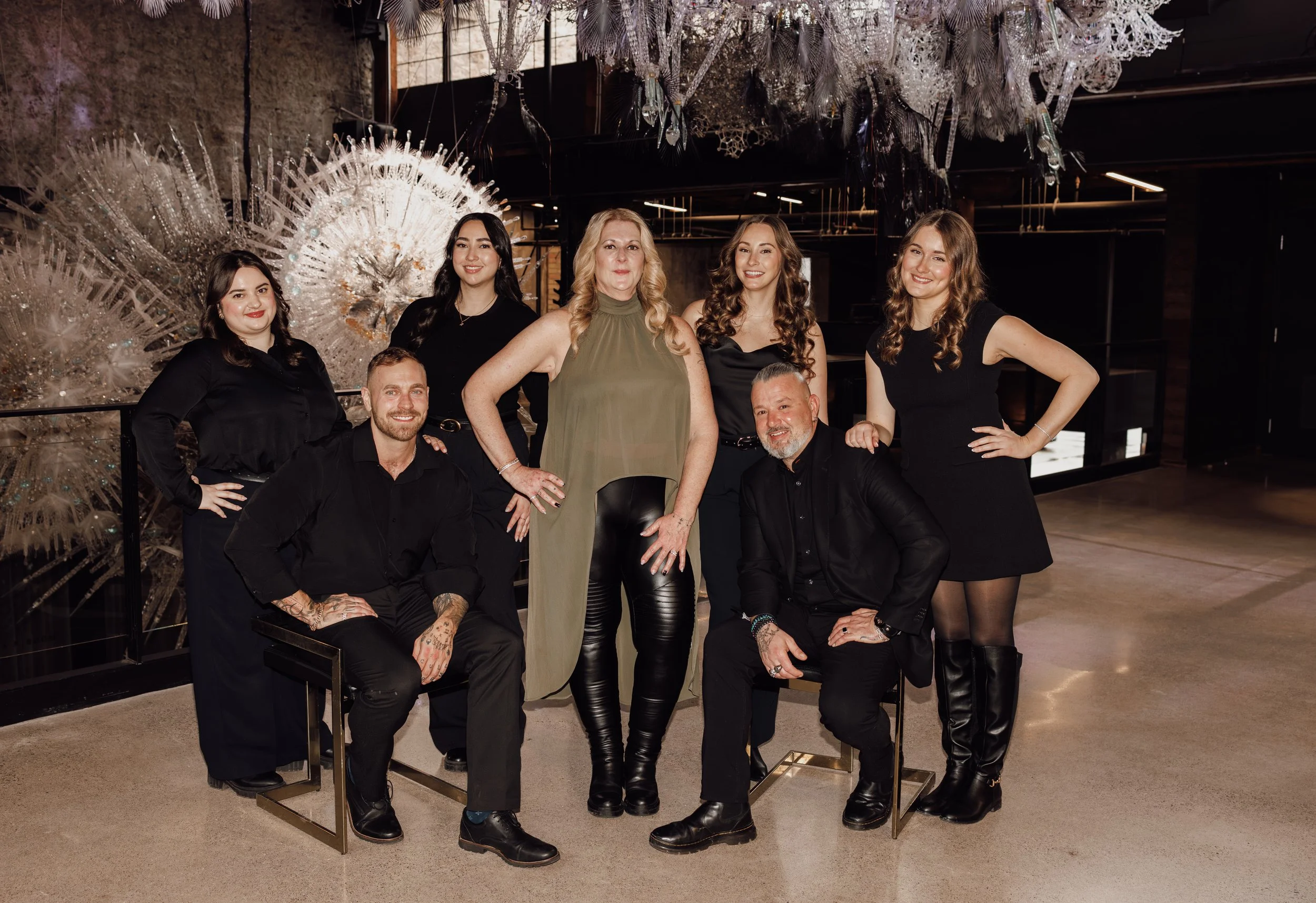 Group of seven smiling people posing for a photo indoors, with decorative art pieces resembling large, intricate, white or clear fan shapes hanging from the ceiling in the background.