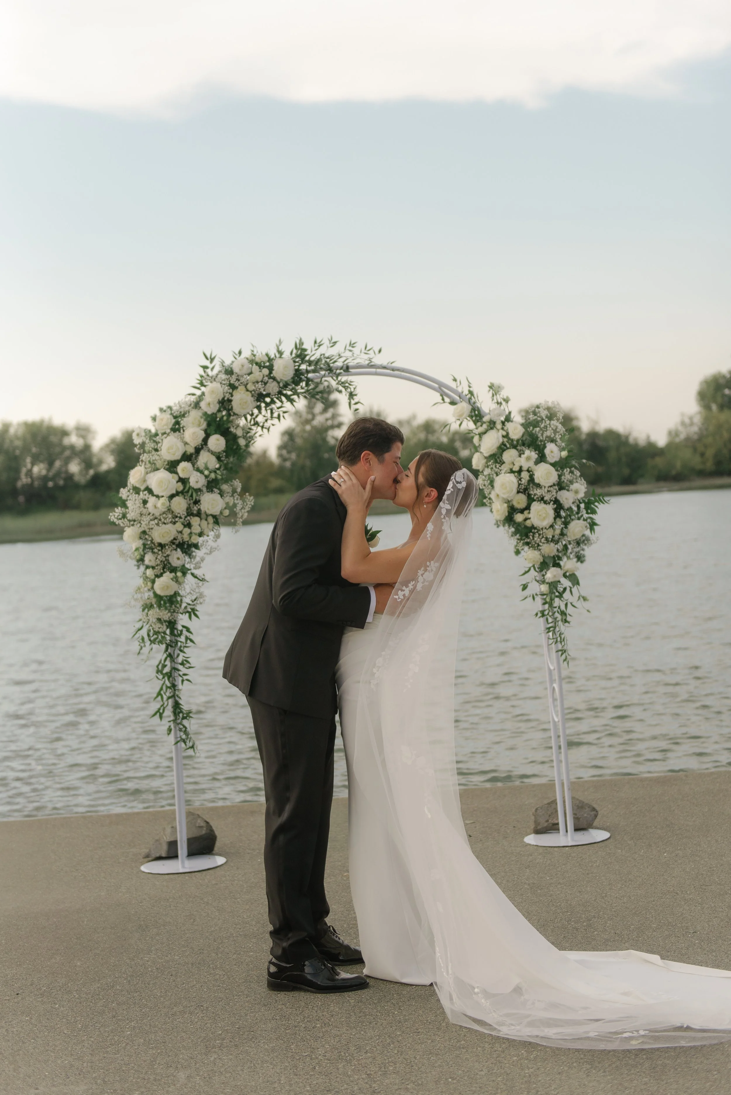 A bride and groom kissing under a floral arch by a body of water during their wedding ceremony.