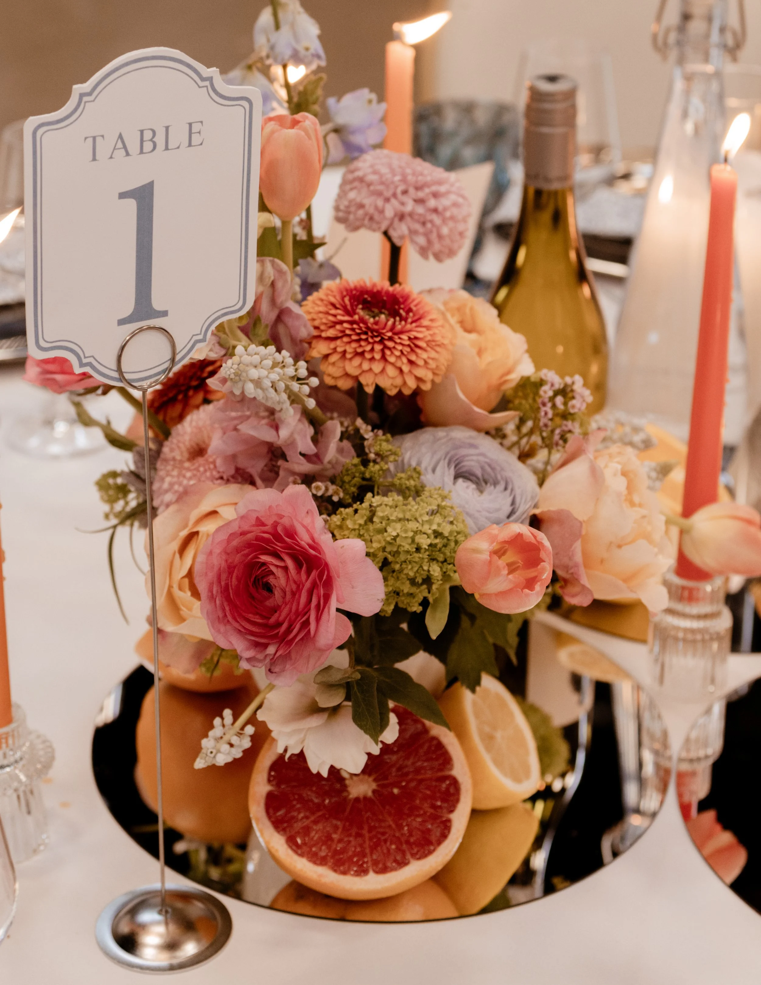 A floral centerpiece with pink, peach, purple, and green flowers on a table, reflected in a mirror. The table has a sign labeled 'Table 1' and sliced grapefruit and other fruits. There are candles and bottles in the background.