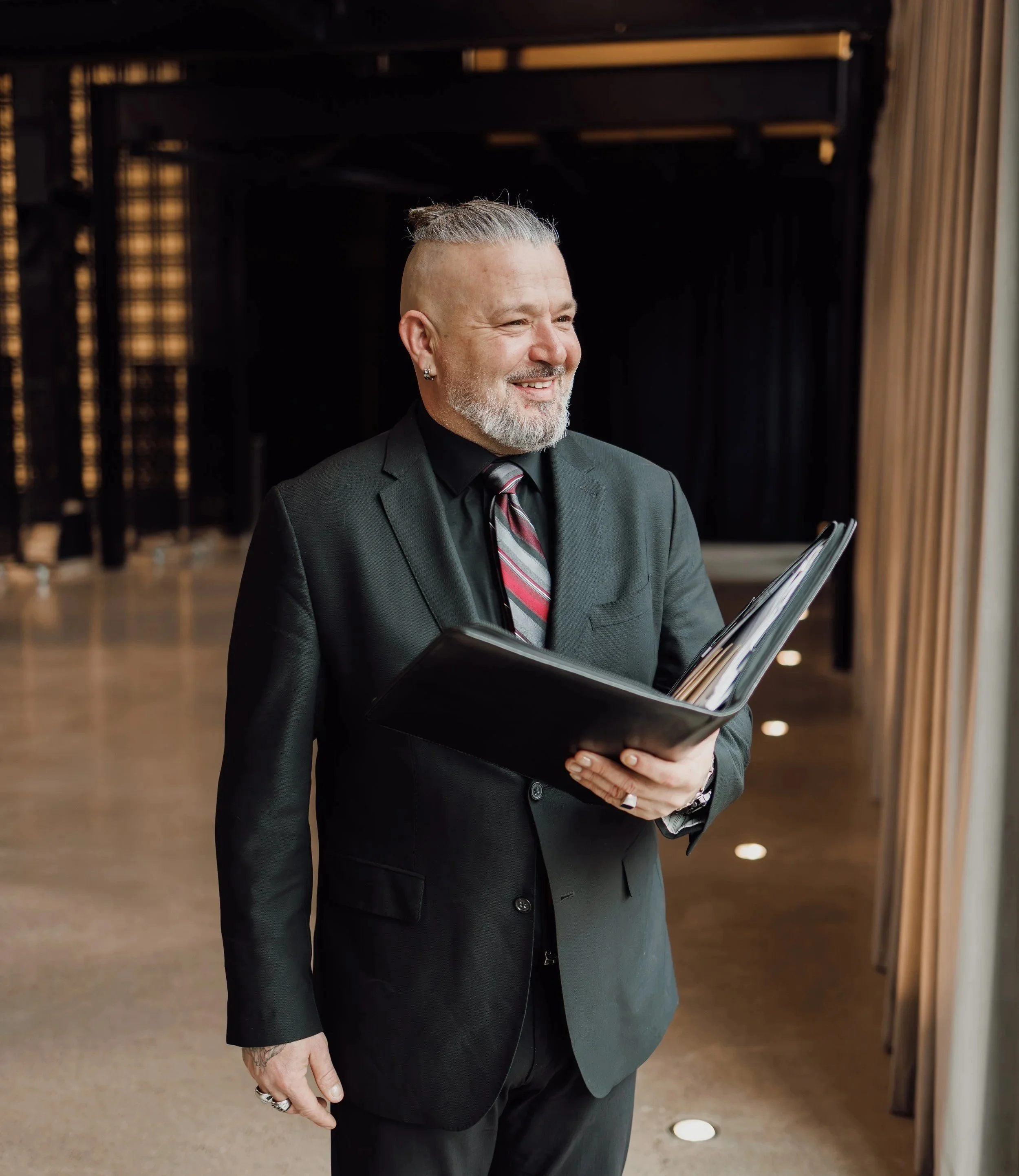 A smiling man dressed in a dark suit with a striped tie, holding a binder, standing in an indoor setting with dark curtains and wooden decor in the background.