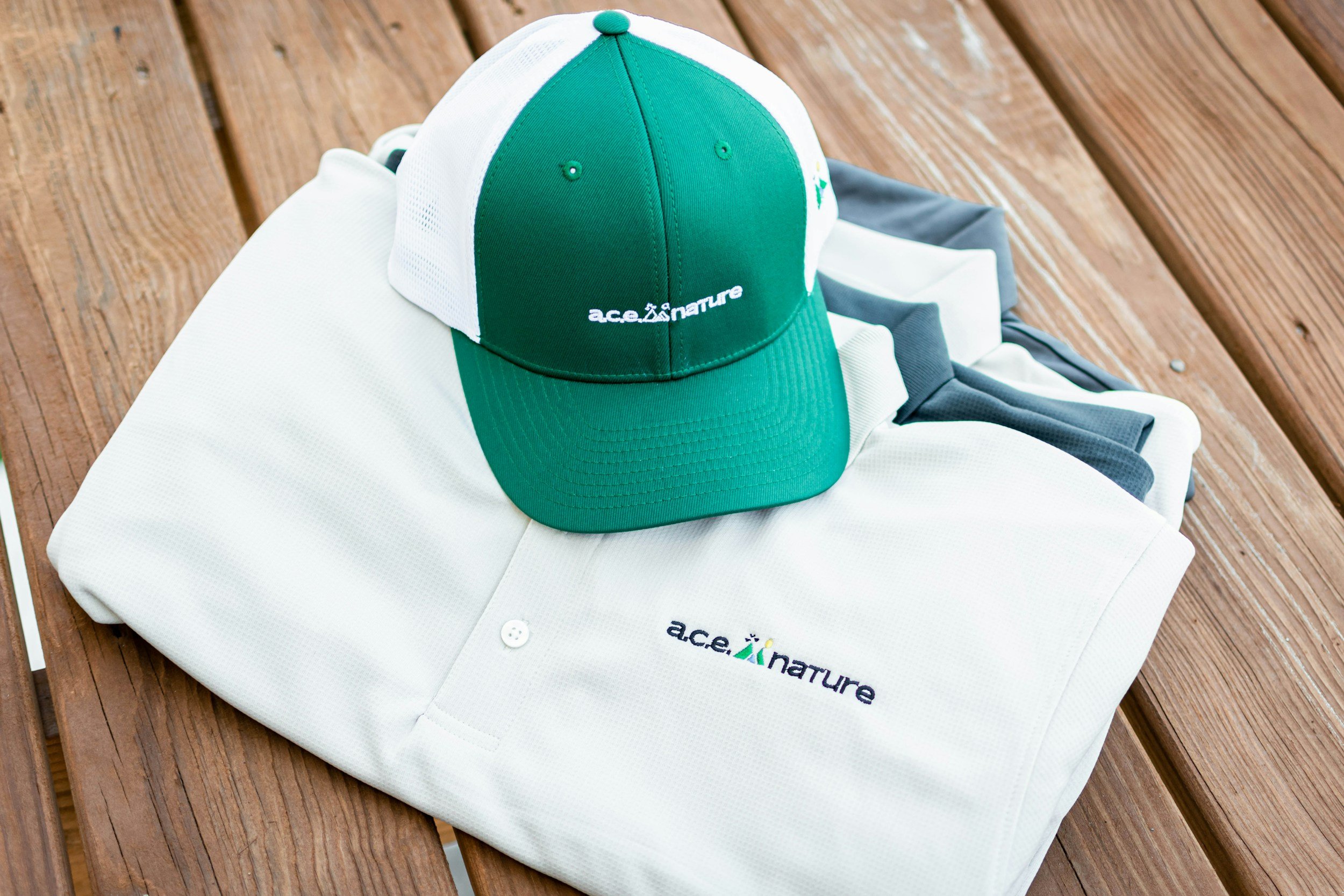 Green and white cap with 'ace of nature' logo on it, resting on a folded white polo shirt with the same logo, on a wooden surface.