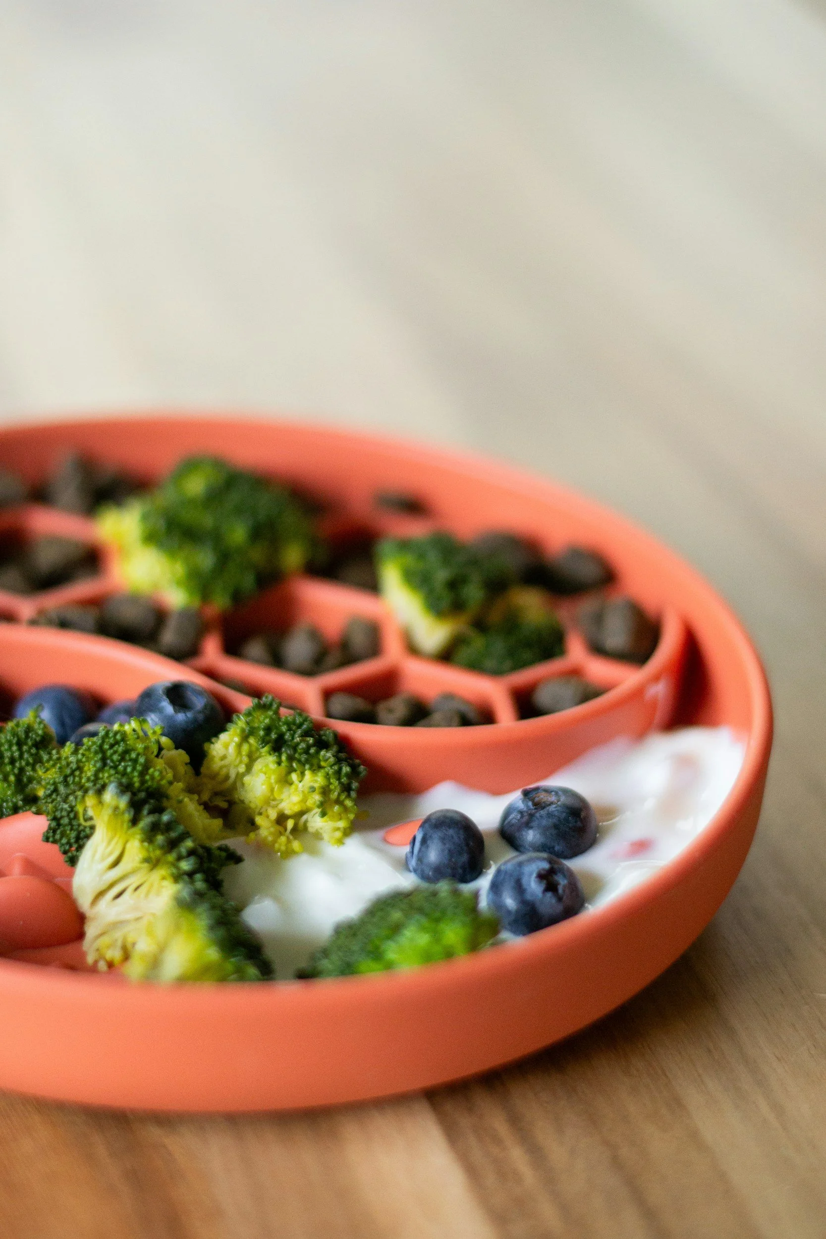 A pink, honeycomb-patterned bowl filled with broccoli, blueberries, and creamy yogurt on a wooden surface.
