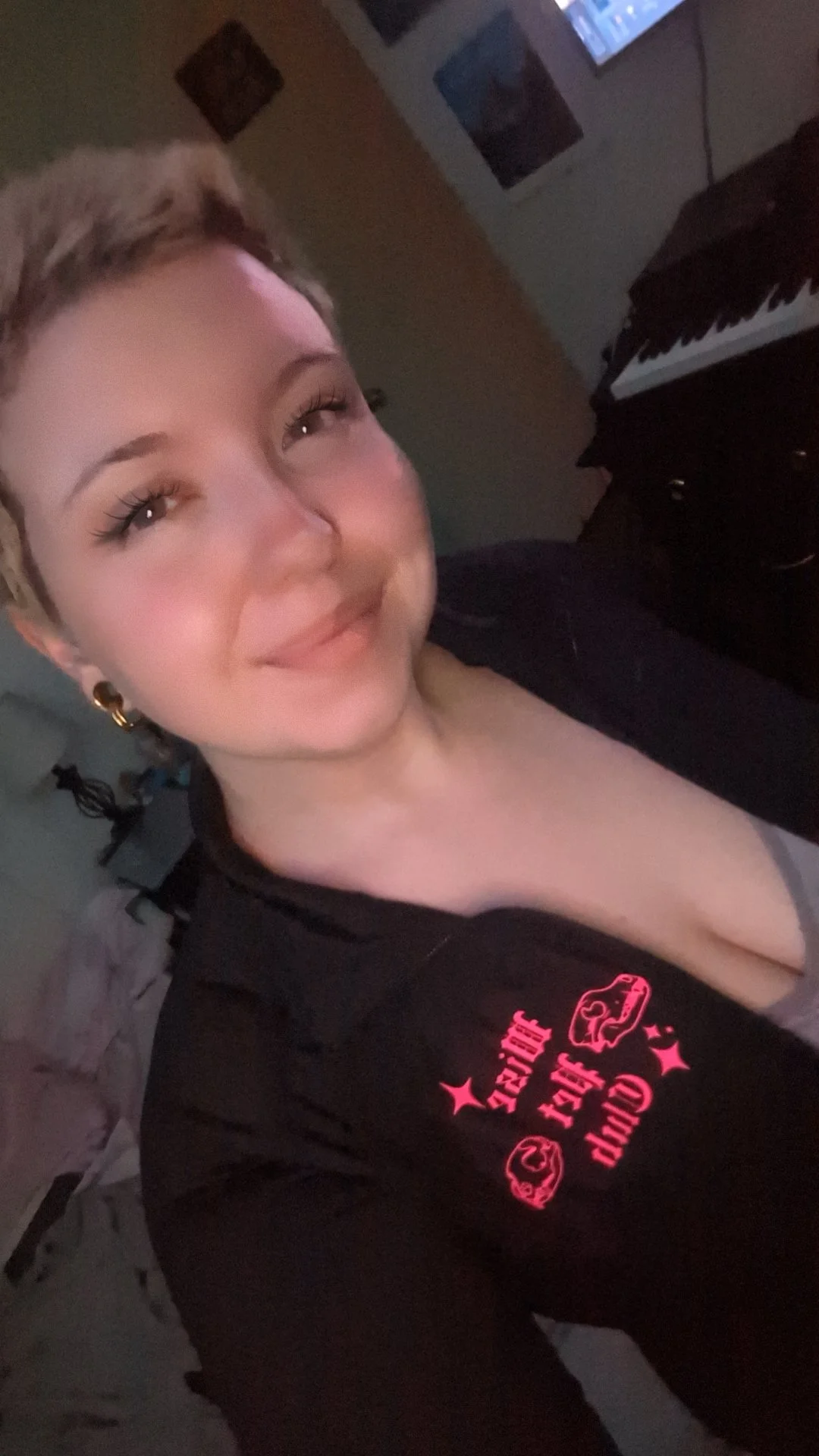 A woman with short curly hair, wearing gold hoop earrings and a black shirt with red graphics, smiling for a selfie in a room with a piano and television in the background.
