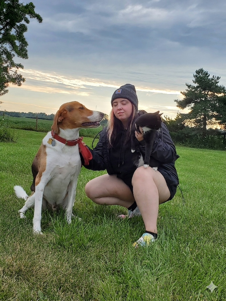 A young woman with light skin, wearing a knit beanie, black jacket, and shorts, is crouched on the grass holding a black and white kitten. Next to her is a large brown and white dog with a red collar and tags, looking at the kitten. The setting is outdoors in a grassy field with trees and a cloudy sky at sunset.