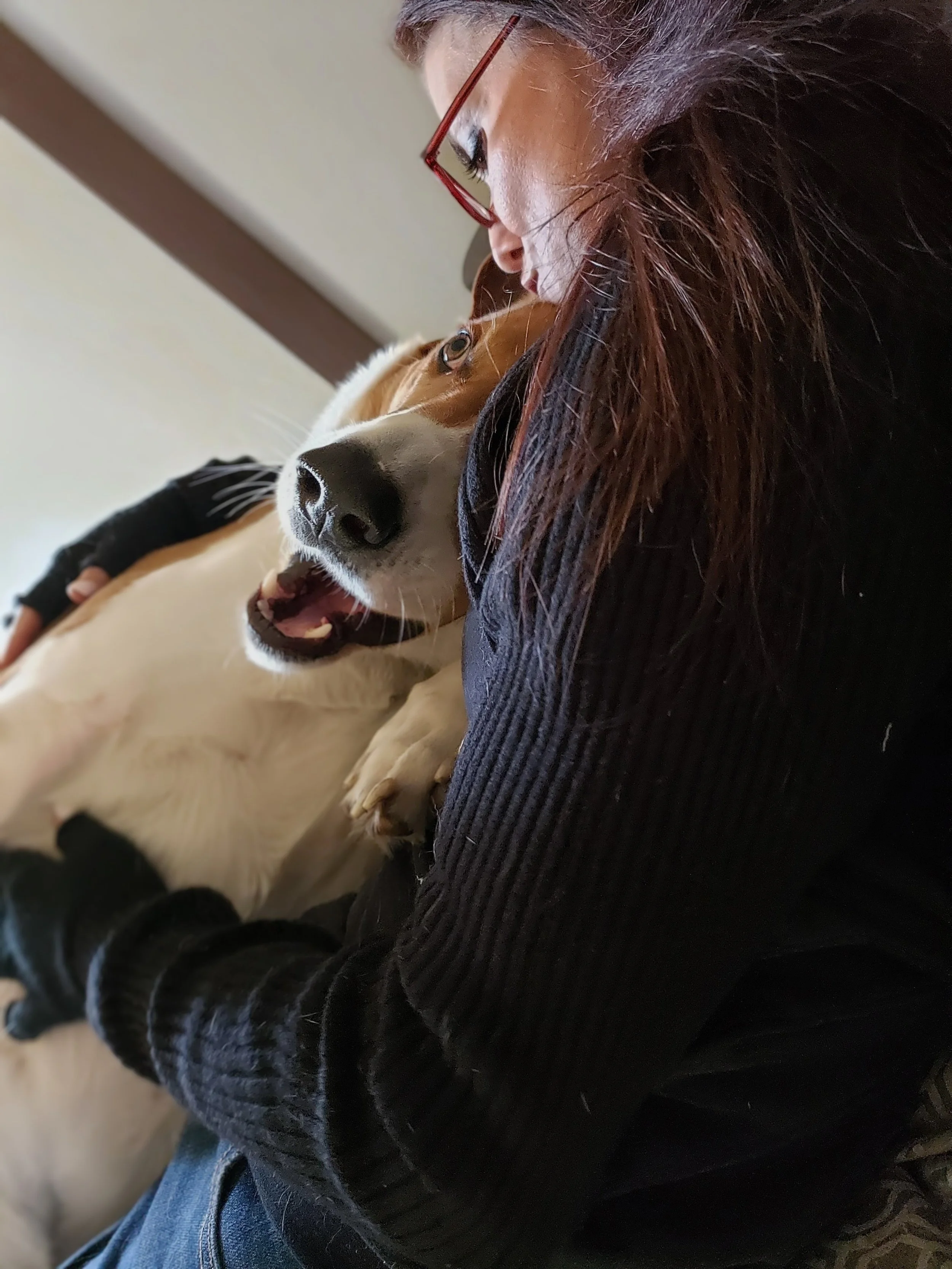 A woman with red glasses and long brown hair is lying close to a light-colored dog with brown markings on its face, one eye, and an open mouth. The dog appears to be relaxed and happy.