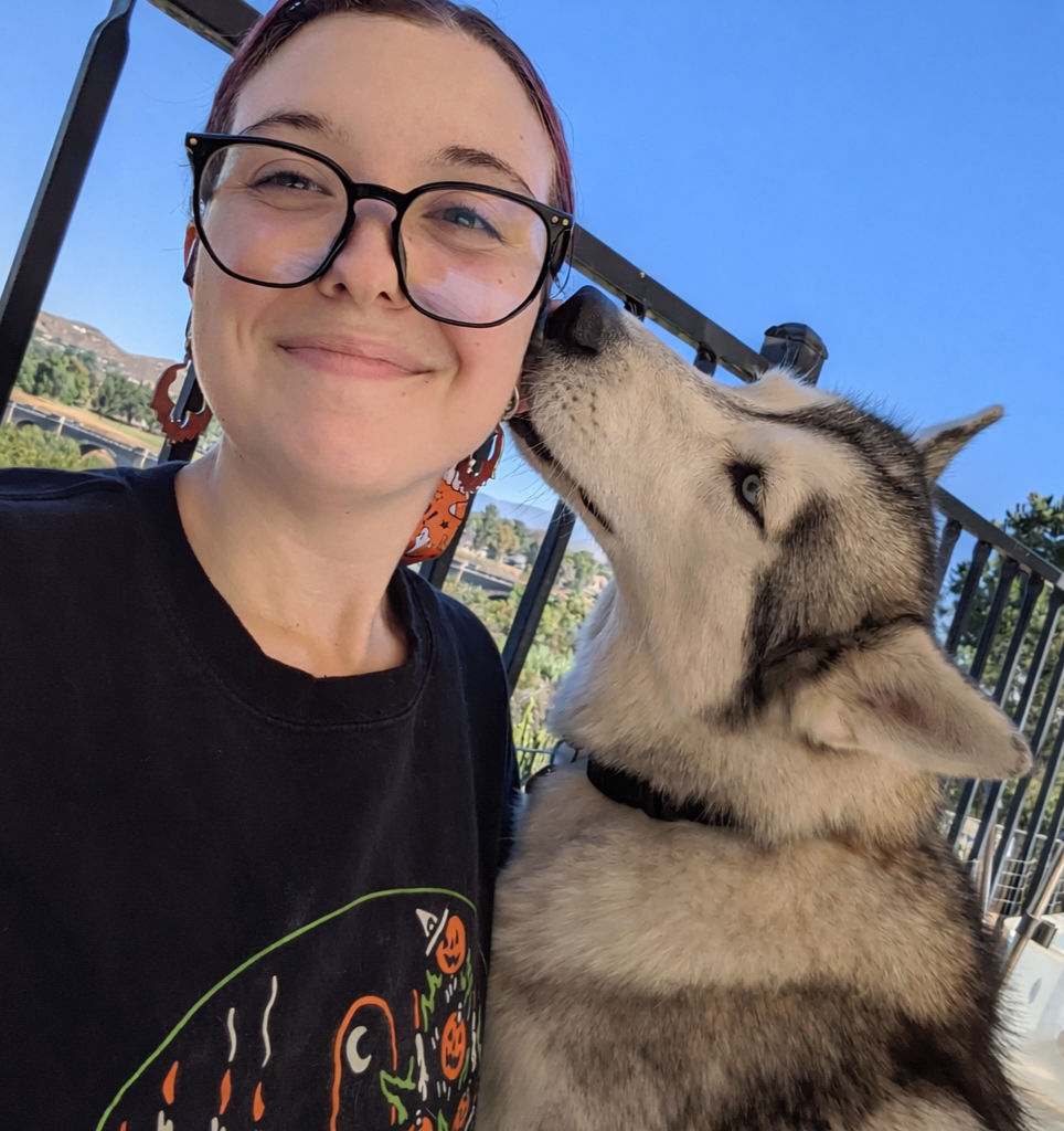 A young woman with glasses and short hair smiling as a husky dog licks her cheek.