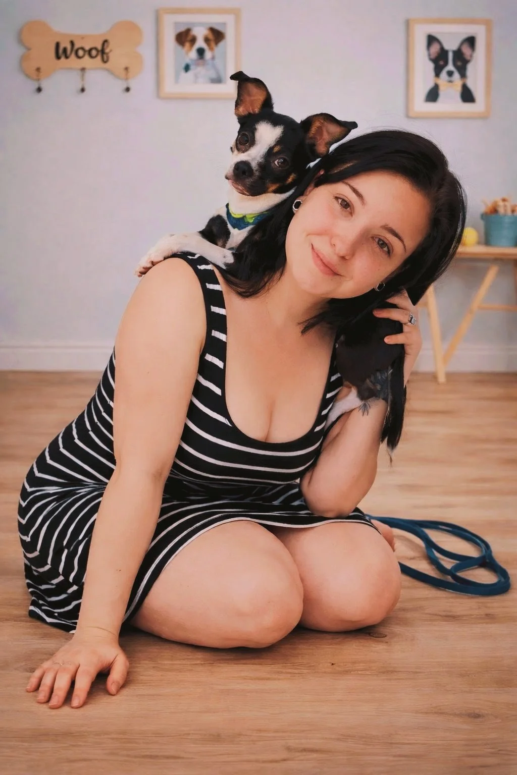 A woman with black hair and a striped black and white dress sitting on the wooden floor, with a small black, white, and tan dog on her shoulder. The woman is smiling and posing for the camera in a room with framed dog pictures and a wall sign that says 'Woof' in the background.