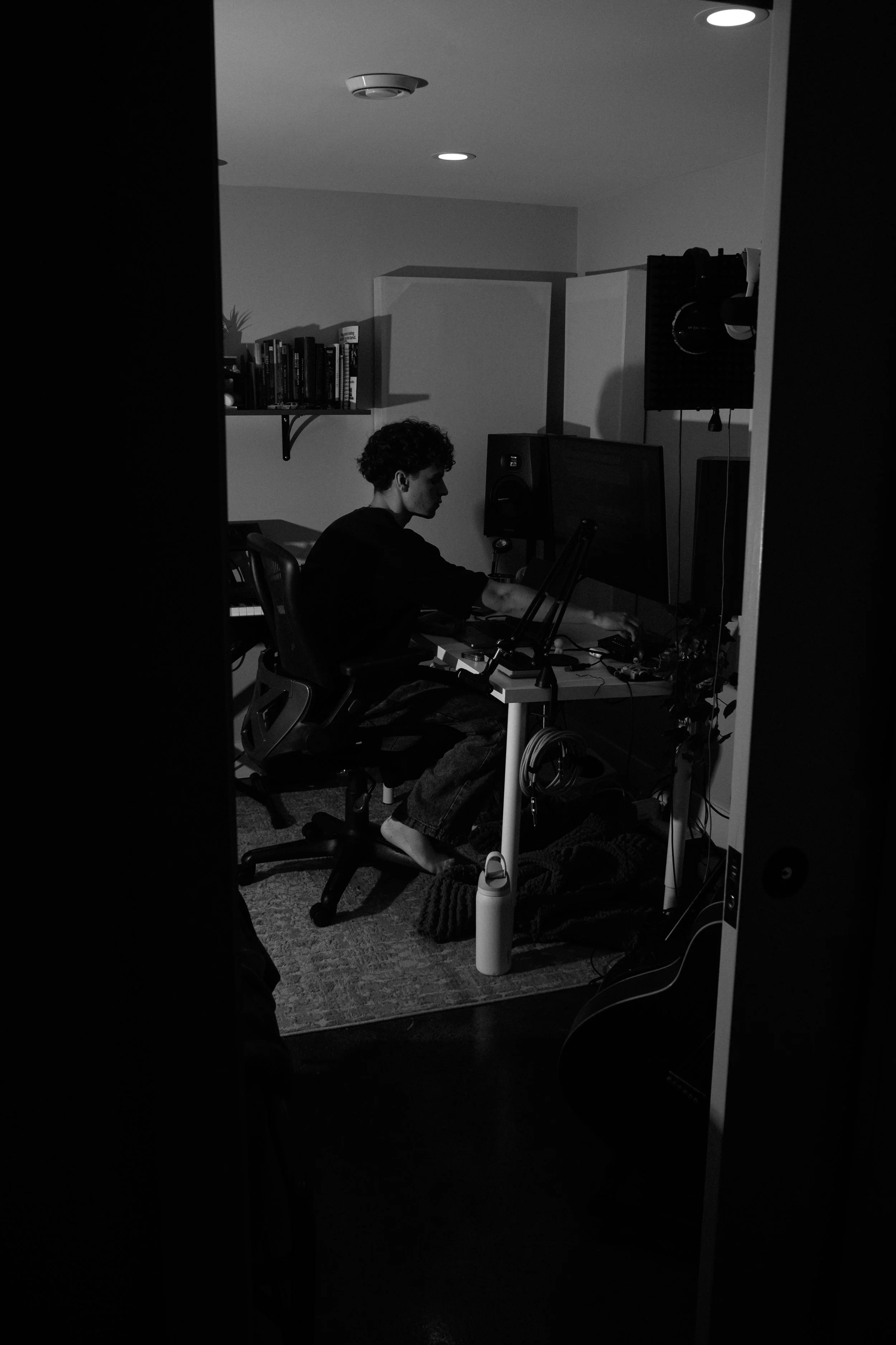 Black and white photo of a person with curly hair sitting at a desk using a computer in a small room. The room has a bookshelf on the wall, a sofa or rug on the floor, and various electronic equipment and cables visible.