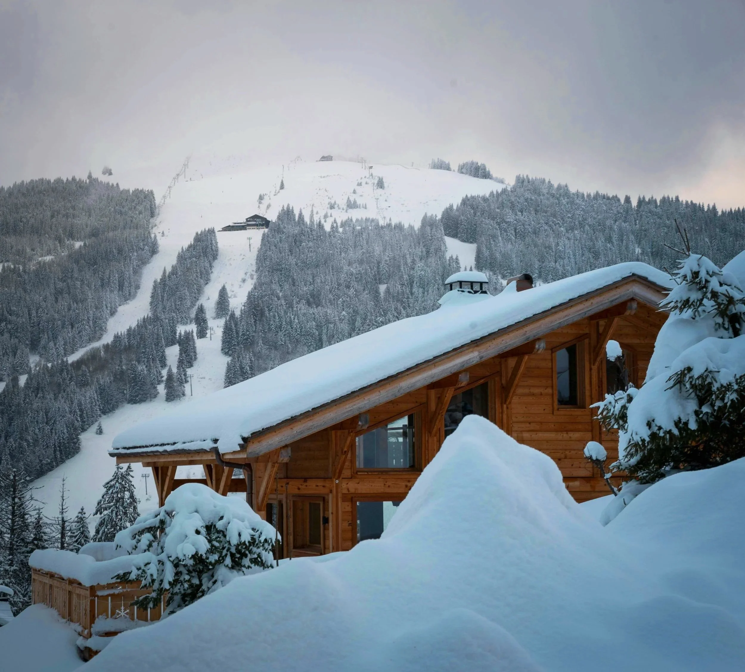 A snowy mountain landscape with a wooden house in the foreground and snow-covered trees. Snow blankets the ground and rooftops, with ski lifts and a ski slope visible on the mountain in the background.