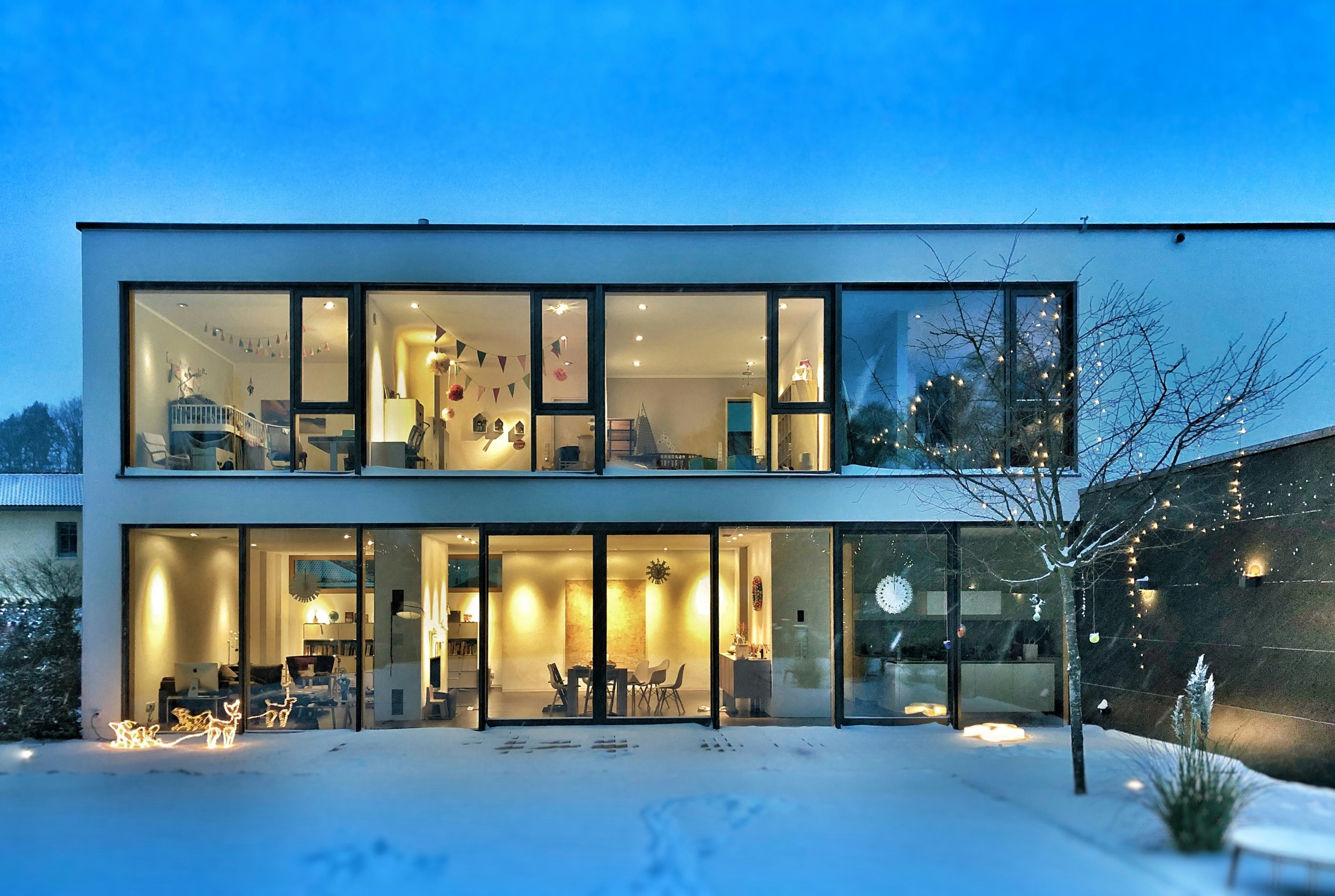 Modern multi-story house with large glass windows illuminated from inside, decorated with holiday lights and ornaments, snow on the ground, and a leafless tree outside during evening.
