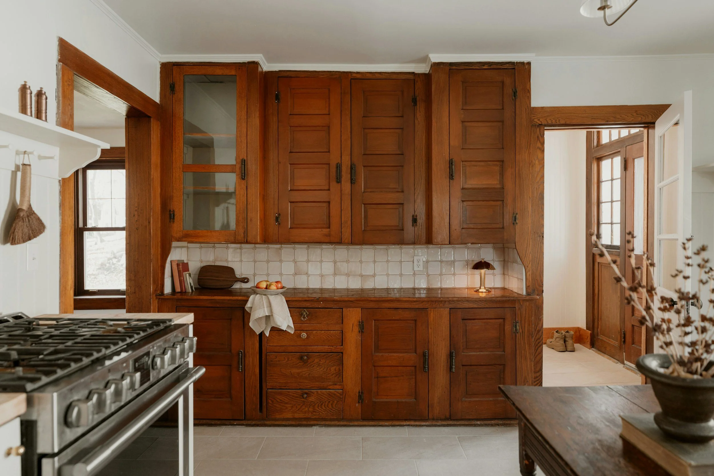 Wooden kitchen cabinets with glass doors above a tiled backsplash, with a window to the side, and a countertop holding a cutting board and fruit bowl.