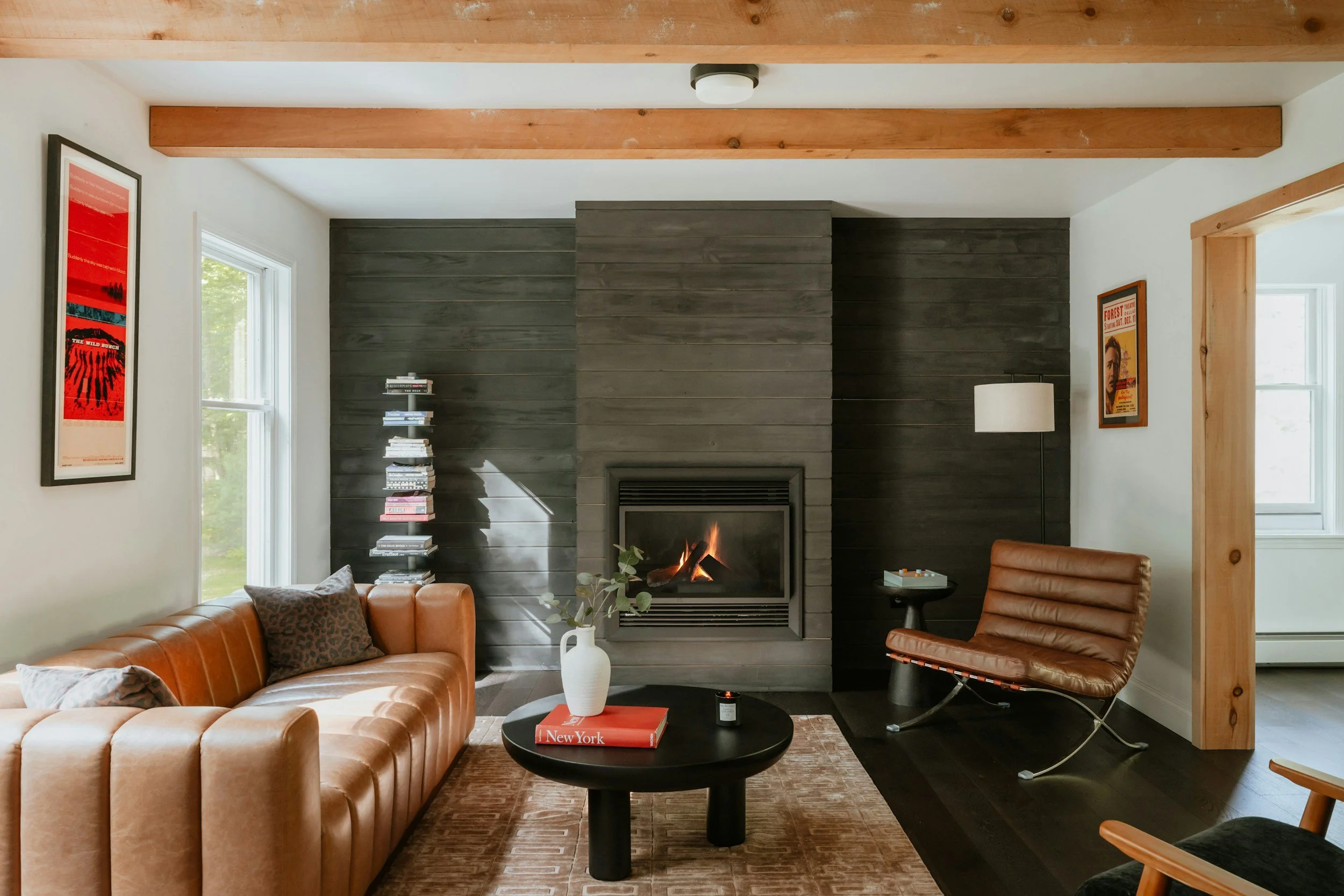 Living room with a fireplace, brown leather sofa, black coffee table with a red book and white vase, and a wooden chair.
