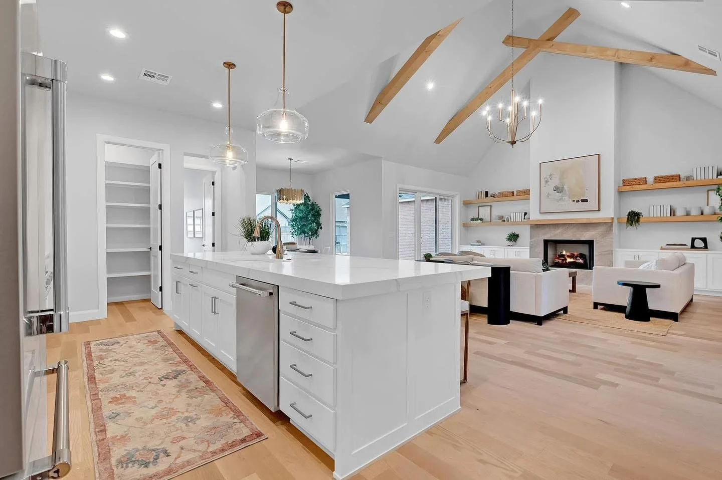 Open-concept modern kitchen and living room with white cabinetry, wooden beams, and a fireplace.