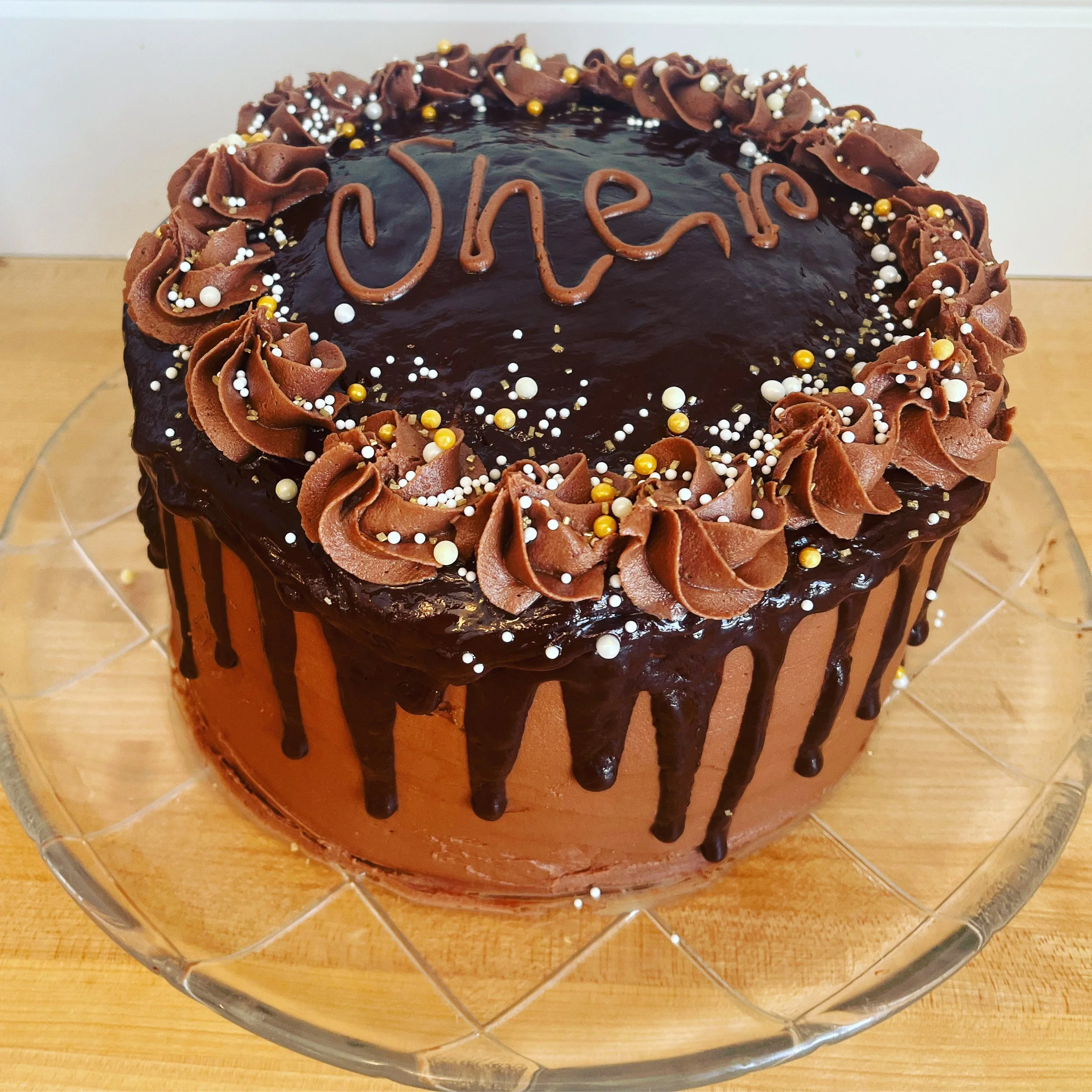 A round chocolate cake with a glossy chocolate glaze, decorated with chocolate frosting swirls, white, gold, and black spherical sprinkles, and the name 'Olea!' written in chocolate on top, in a glass cake stand.