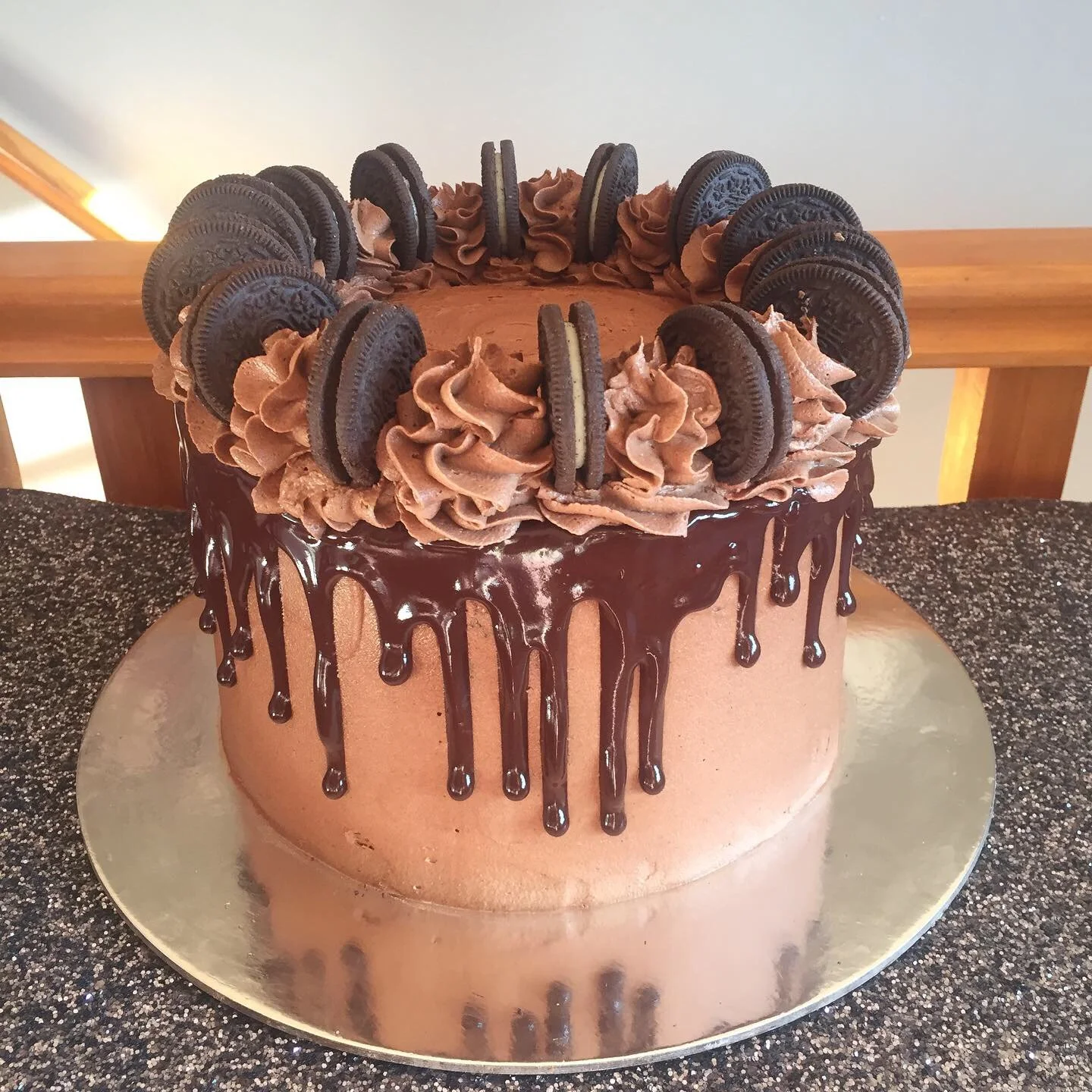 Chocolate cake with Oreo cookies and chocolate frosting on top, with chocolate drizzle over the sides, on a round silver cake board.