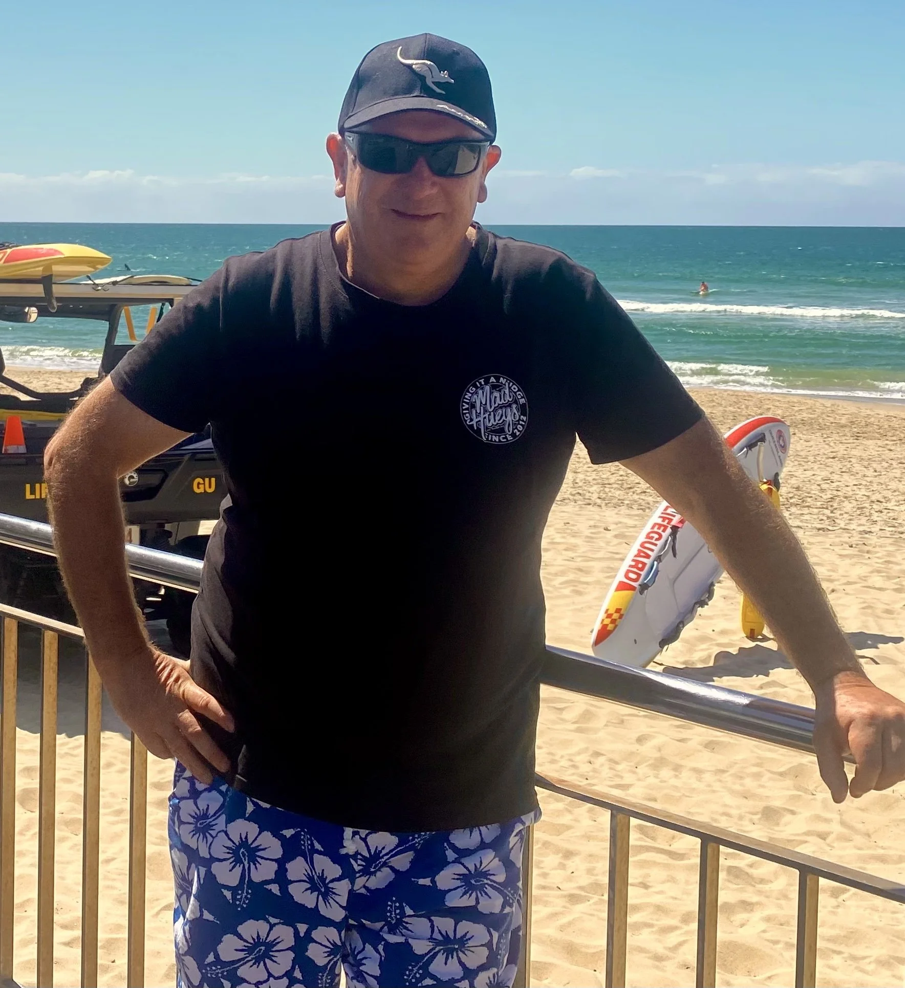 Grant Clifton, Financial Adviser, standing on a beach in Queensland. The background shows the ocean with a person surfing and a sandy shoreline.