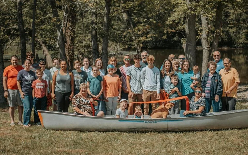 Group of people, including children and adults, gathered outdoors near a river with trees, some sitting in a canoe and others standing behind it, celebrating an event with a ribbon, during daytime.