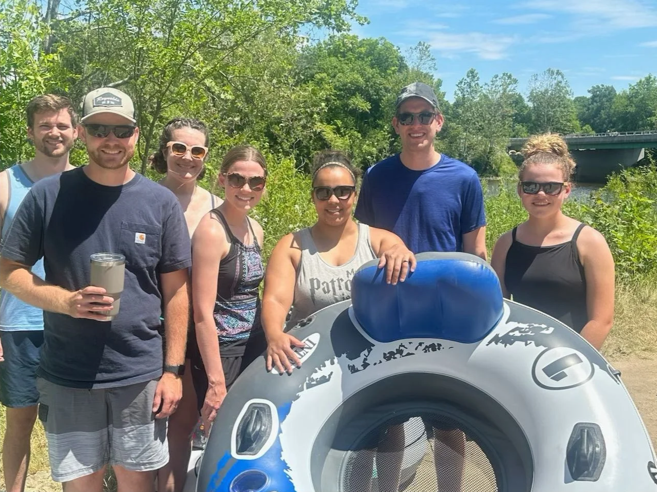Group of eight people outdoors next to river, posing for photo with a paddleboard.