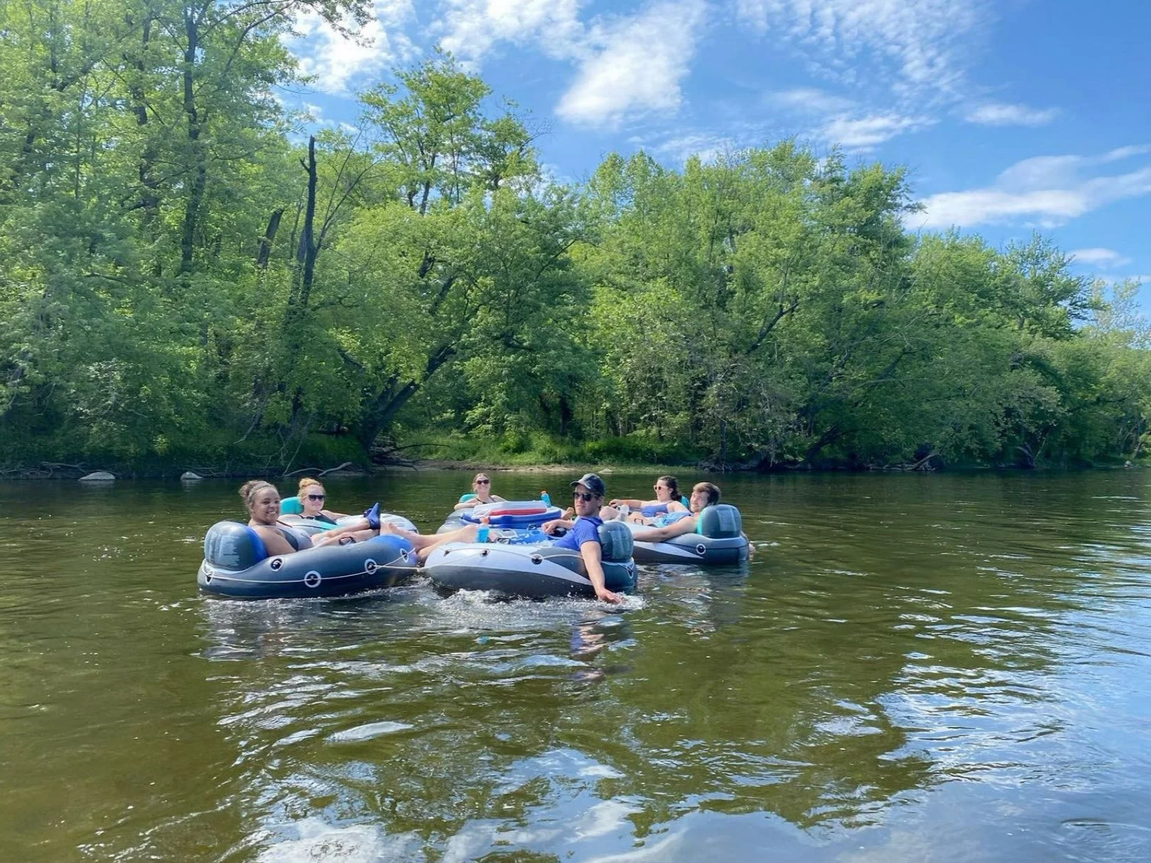 Six people floating on inflatable boats in a river surrounded by green trees and blue sky.
