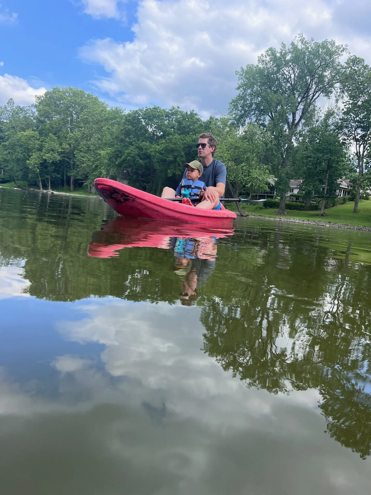 A man and a young child sit in a red kayak on calm water, surrounded by green trees under a partly cloudy sky.