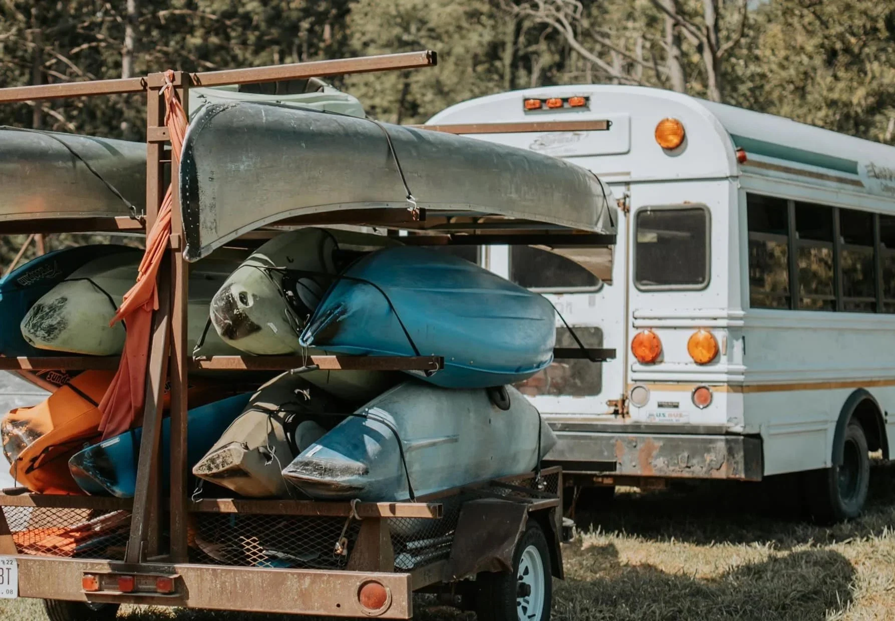 A trailer carrying multiple kayaks on a rusted metal rack attached to a rusty truck with an older design, parked outdoors with trees in the background.