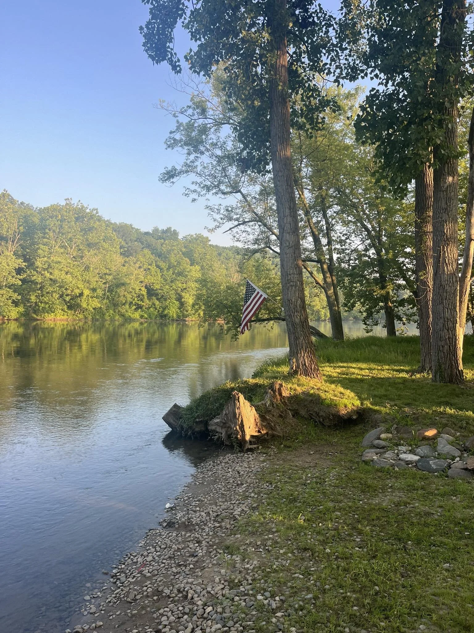 A peaceful lakeside scene with trees along the shore, reflecting in the water, with a small American flag hanging from one of the trees.