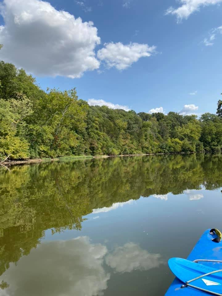 Calm river with lush green trees along the banks, blue sky with scattered clouds, and a blue kayak partially visible in the lower right corner, reflecting the scenery in the water.