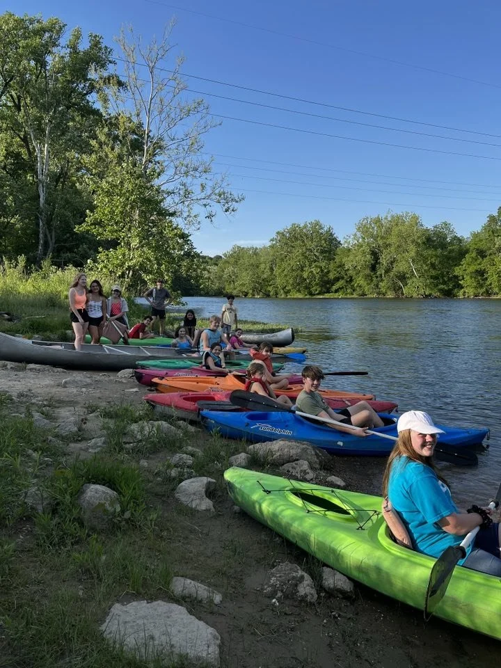 Group of children and teenagers in colorful kayaks and canoes on a riverbank surrounded by trees on a sunny day.