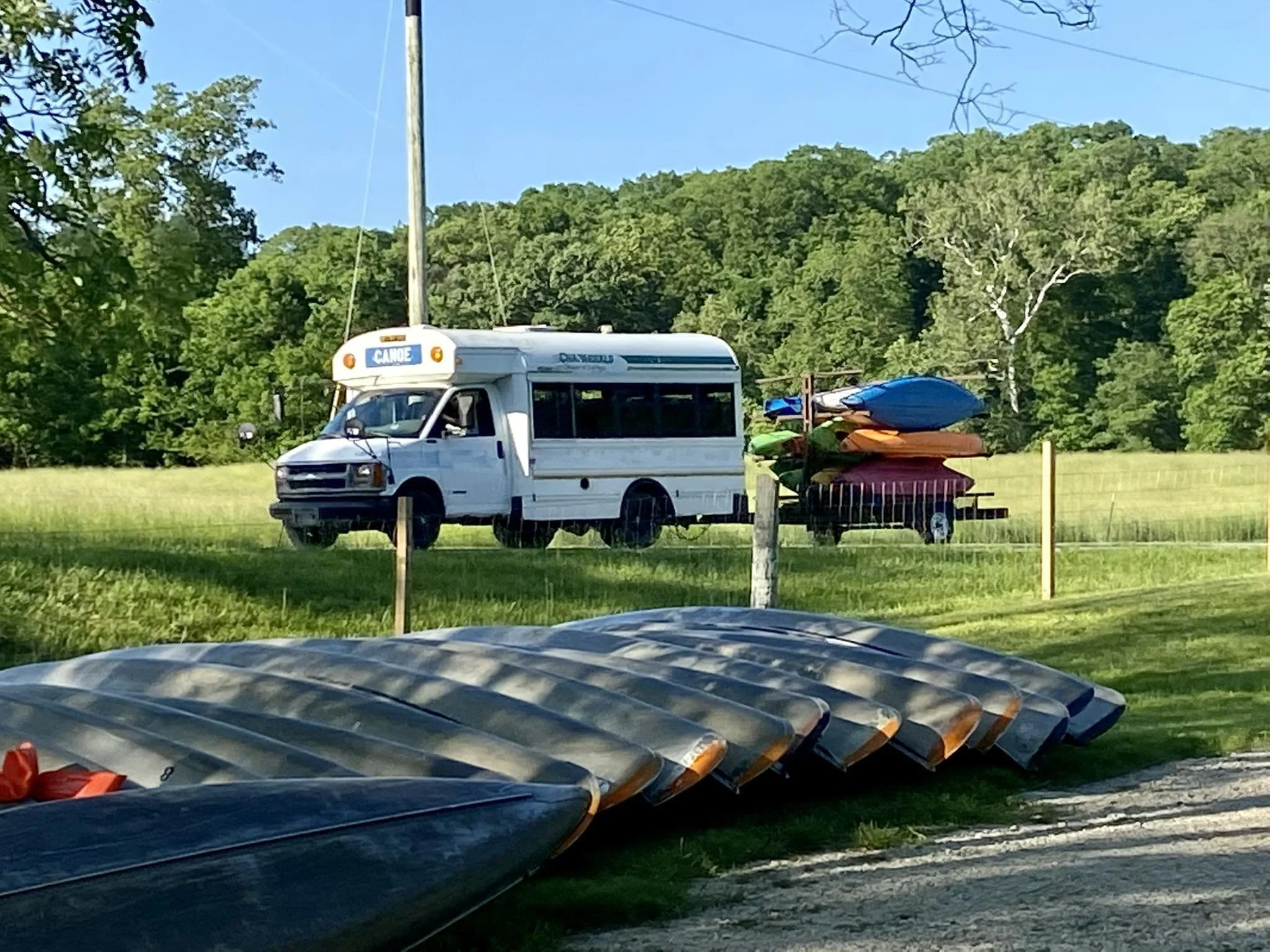 View of a grassy outdoor area with a white camper van labeled 'CAMPER' parked near a wooden fence. Several colorful kayaks are stacked on a cart and placed near the fence. The background includes lush green trees and a blue sky.