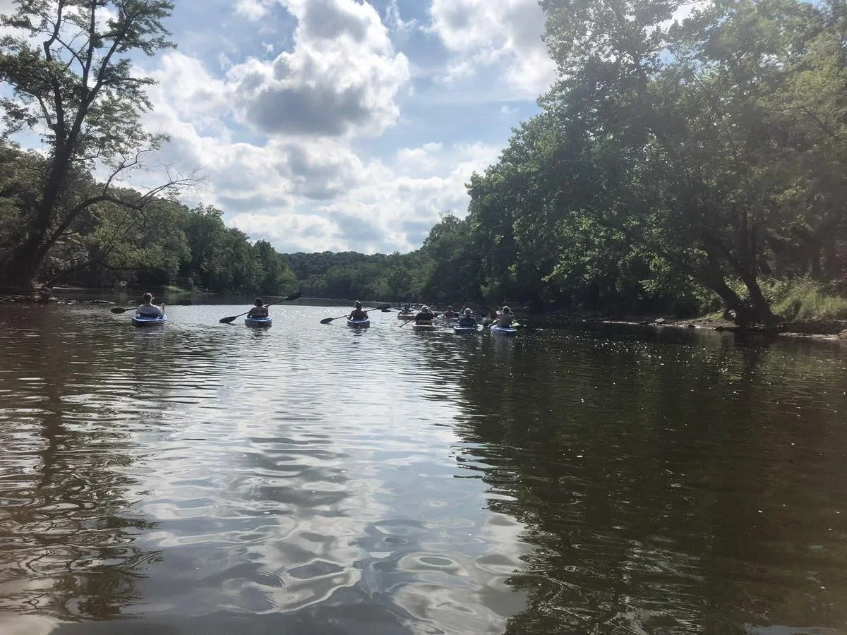 People kayaking on a river surrounded by trees and under partly cloudy sky.