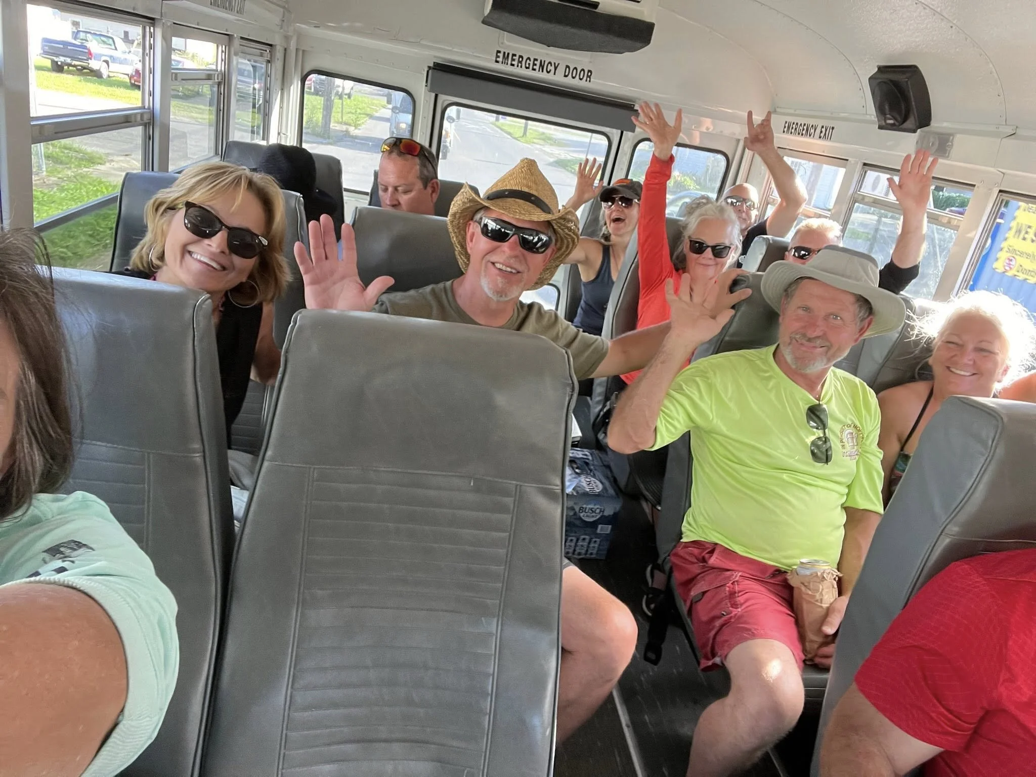 A group of people sitting inside a bus, smiling and waving at the camera, with some wearing sunglasses, hats, and bright clothing, suggesting they are on a fun outing or tour.