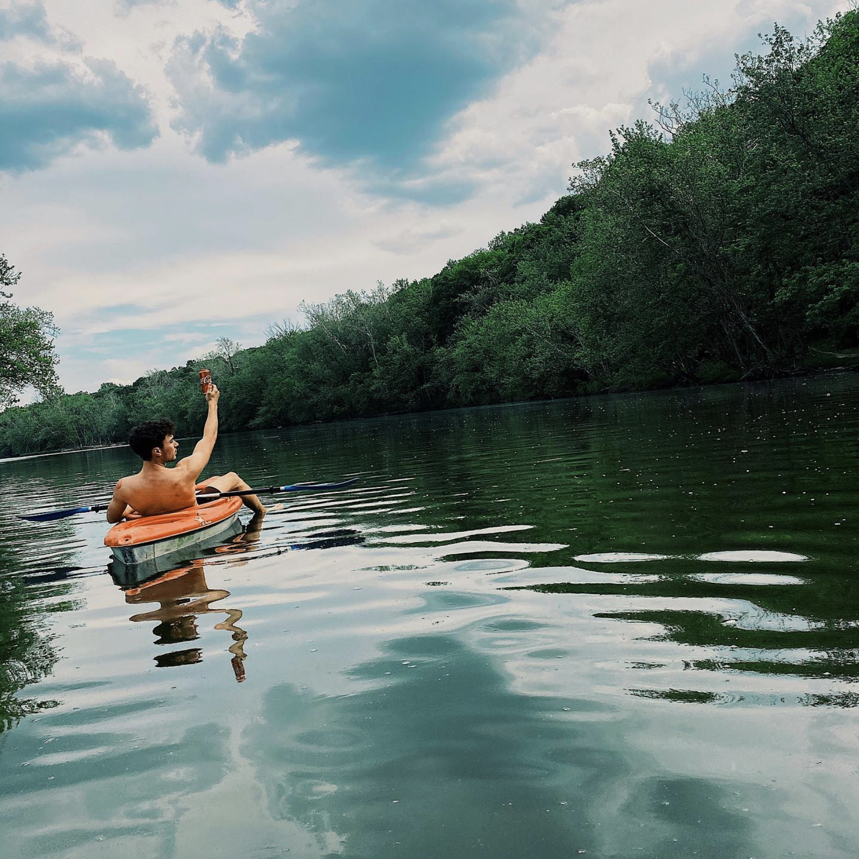 A person on an orange kayak holding a Coca Cola can while on a body of water surrounded by trees and a cloudy sky.