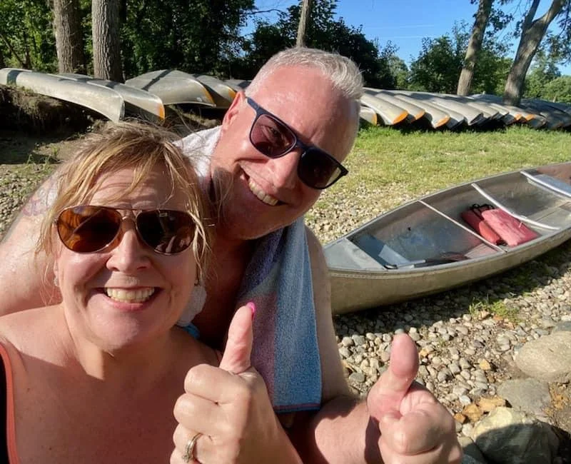 A smiling woman and man wearing sunglasses, giving thumbs up in front of parked canoes and kayaks near a lake or river.