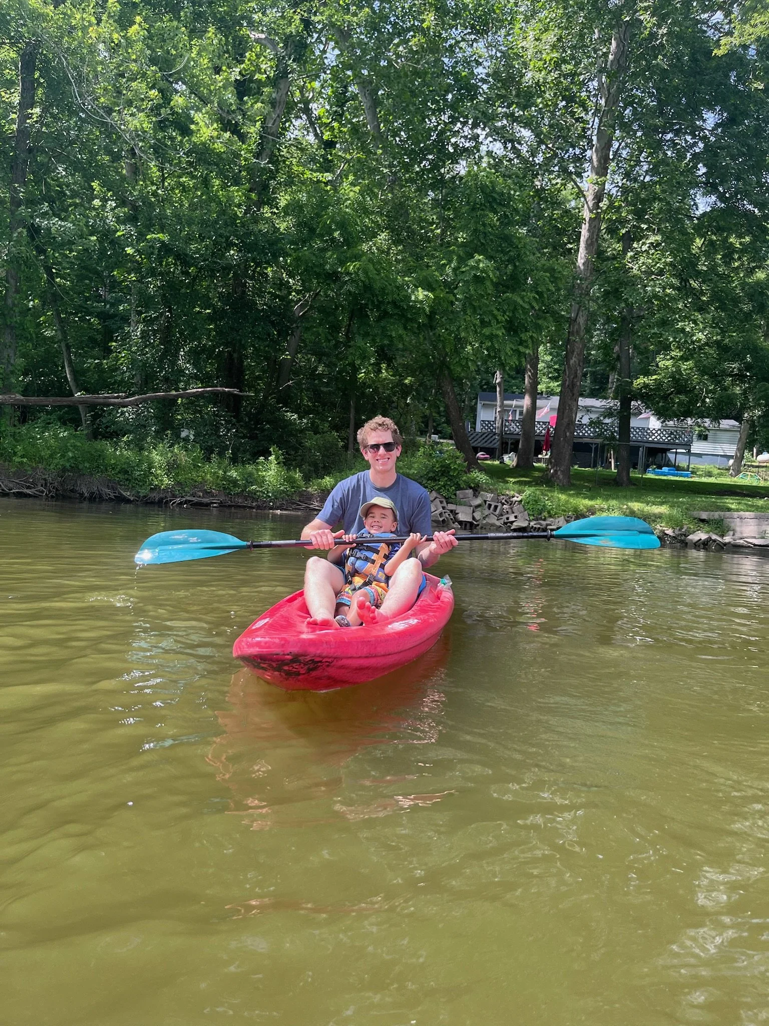 A man and a child kayaking on a river with trees and houses in the background.