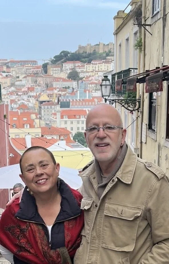 A smiling couple standing outdoors on a hillside with a cityscape in the background, featuring colorful buildings and a castle.