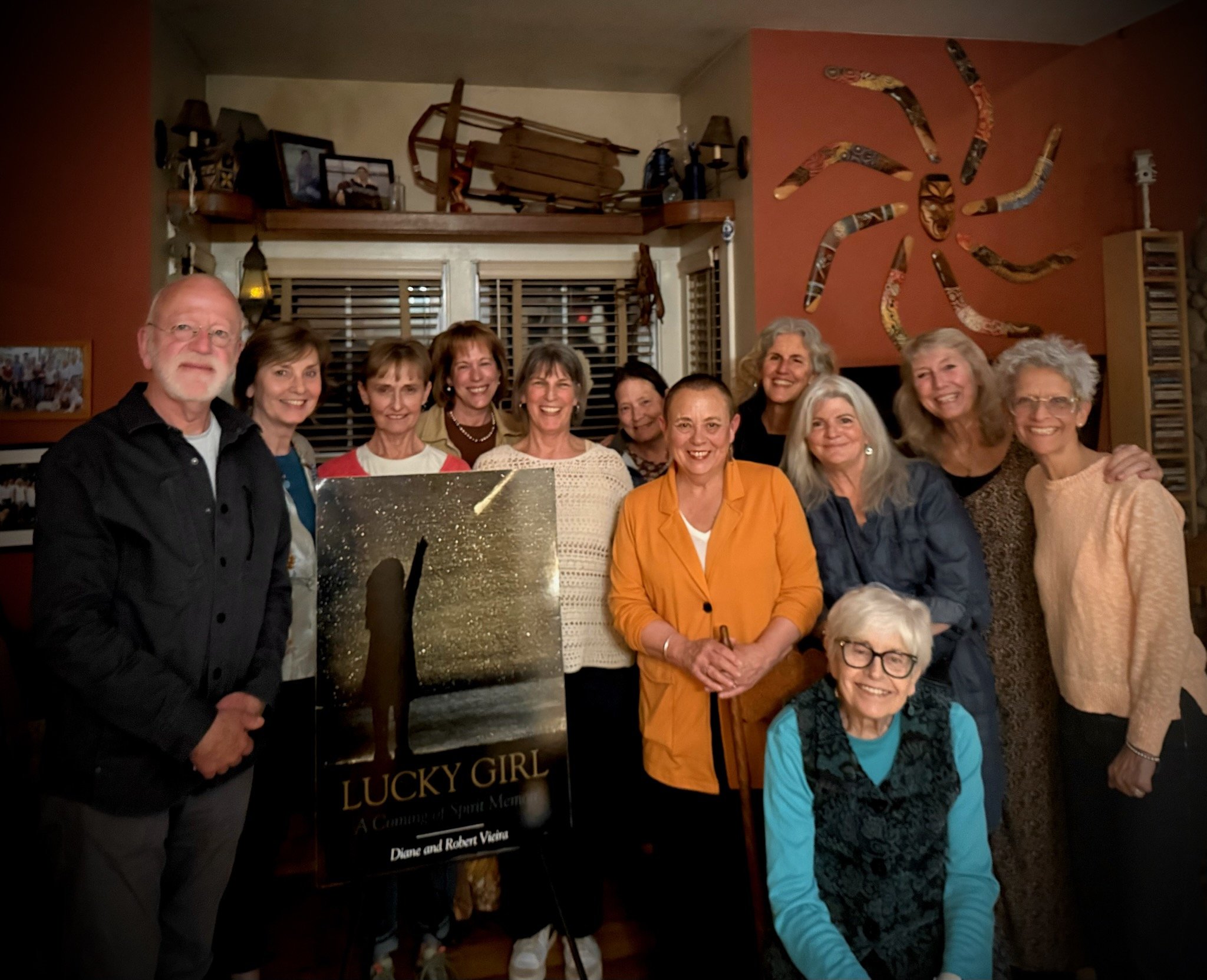A group of people smiling and posing together indoors, with a sign in front of them that reads "Lucky Girl." The background features a decorative wall with a snake and a bow, and shelves with various items.