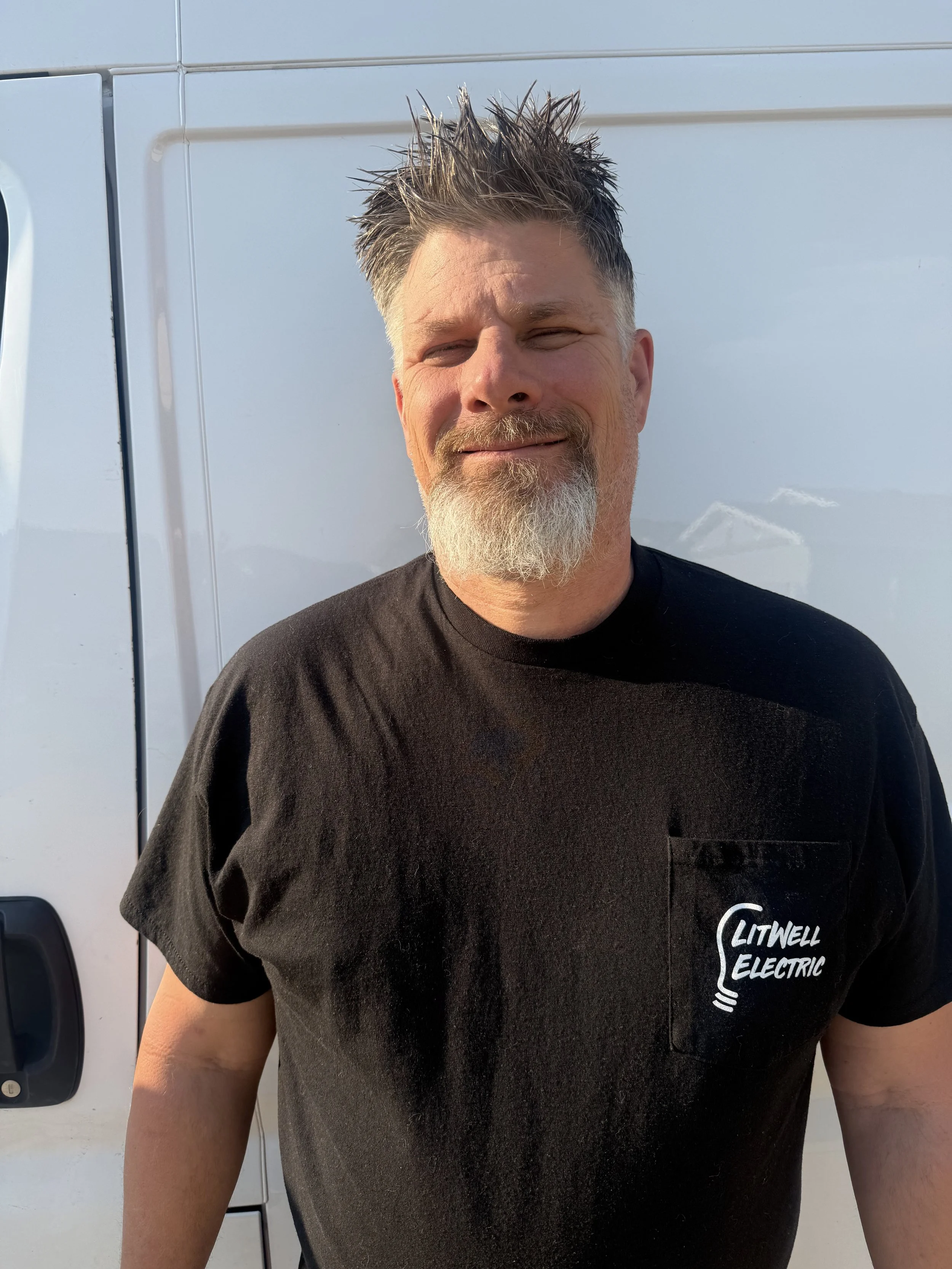A man with a beard and spiky hair smiling while standing in front of a white utility vehicle with a company logo that reads "Litwell Electric."