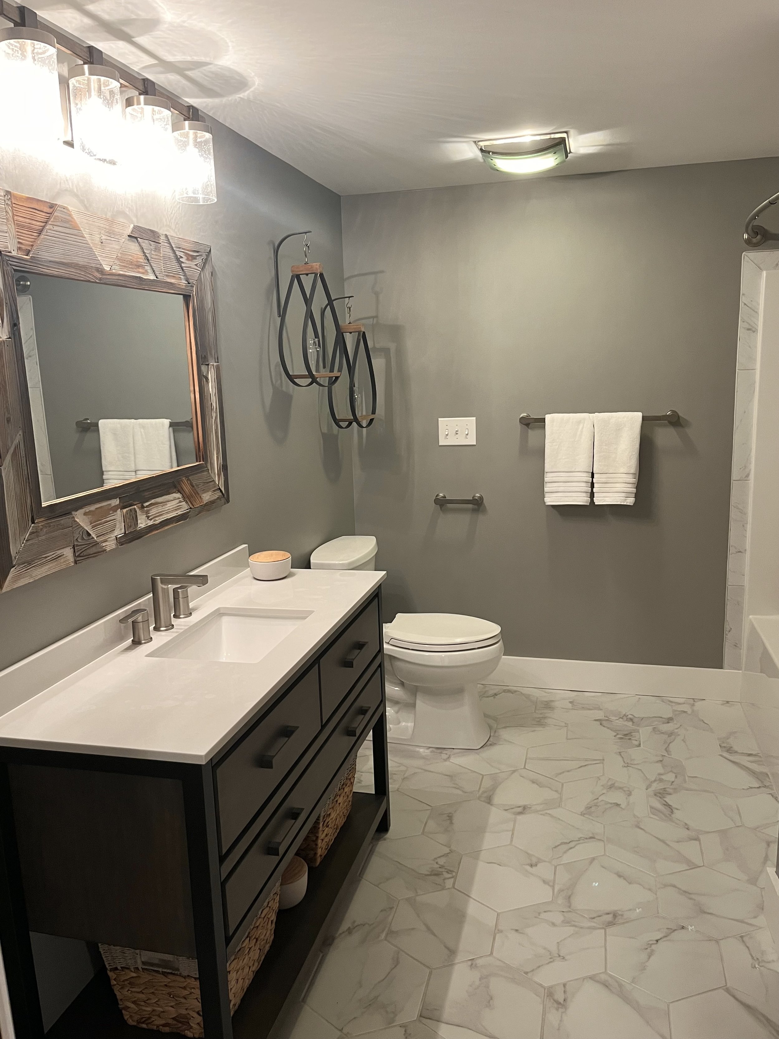 A modern bathroom featuring a white marble hexagon floor, a dark vanity with a white countertop, a rectangular mirror with a rustic wooden frame, wall-mounted towel racks with white towels, decorative black metal wall shelves with glass jars, a toilet, and a shower area with a glass partition. The walls are painted light gray, and the lighting includes a ceiling fixture and vanity lights.