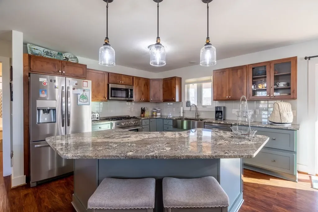 A modern kitchen with a granite island, stainless steel appliances, wooden cabinets, and three hanging pendant lights.