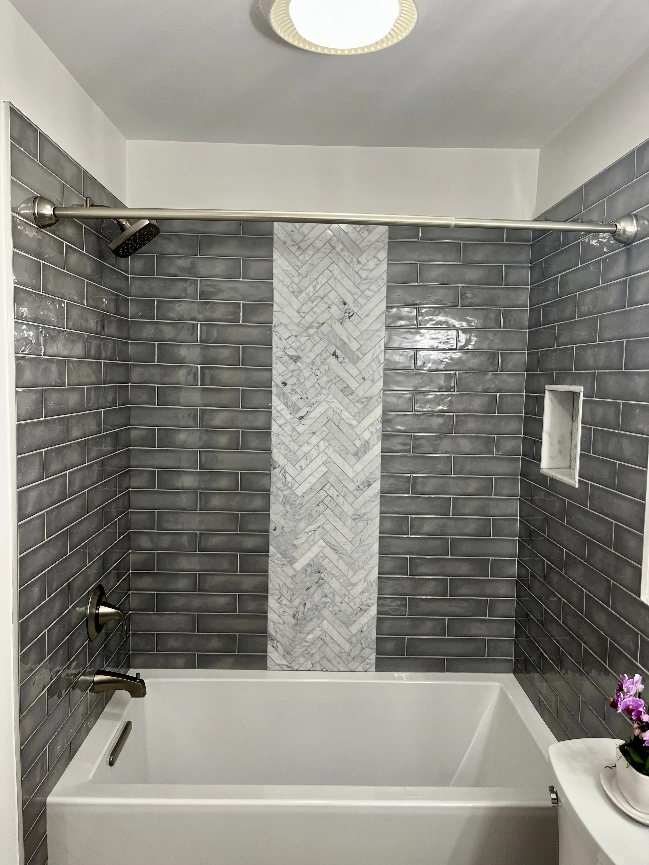 Modern bathtub shower with gray subway tiles, a vertical marble accent strip, a built-in shelf, a orchid plant, and a ceiling light fixture.