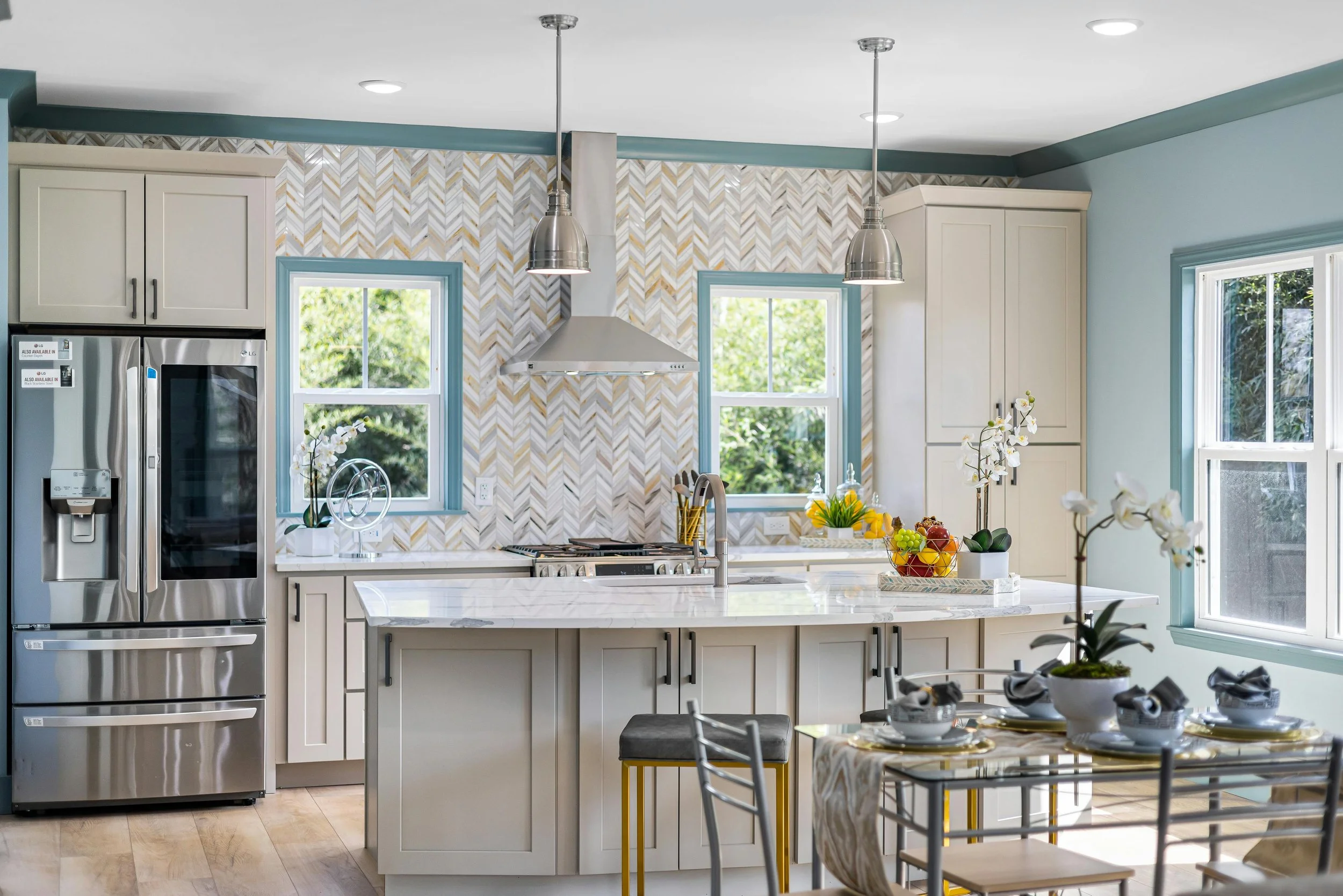 A modern kitchen with white cabinets, a marble island, stainless steel refrigerator, and pendant lighting. There are two windows with views of greenery, and a table with dishes and floral centerpieces.