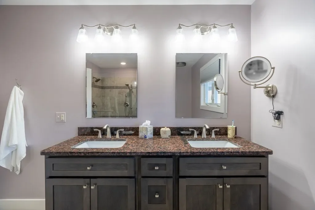 Double vanity bathroom with granite countertop, two sinks, mirrors above, wall-mounted lights, a towel hanging on the left wall, and a wall-mounted magnifying mirror on the right, with a view of a shower in the mirror reflection.