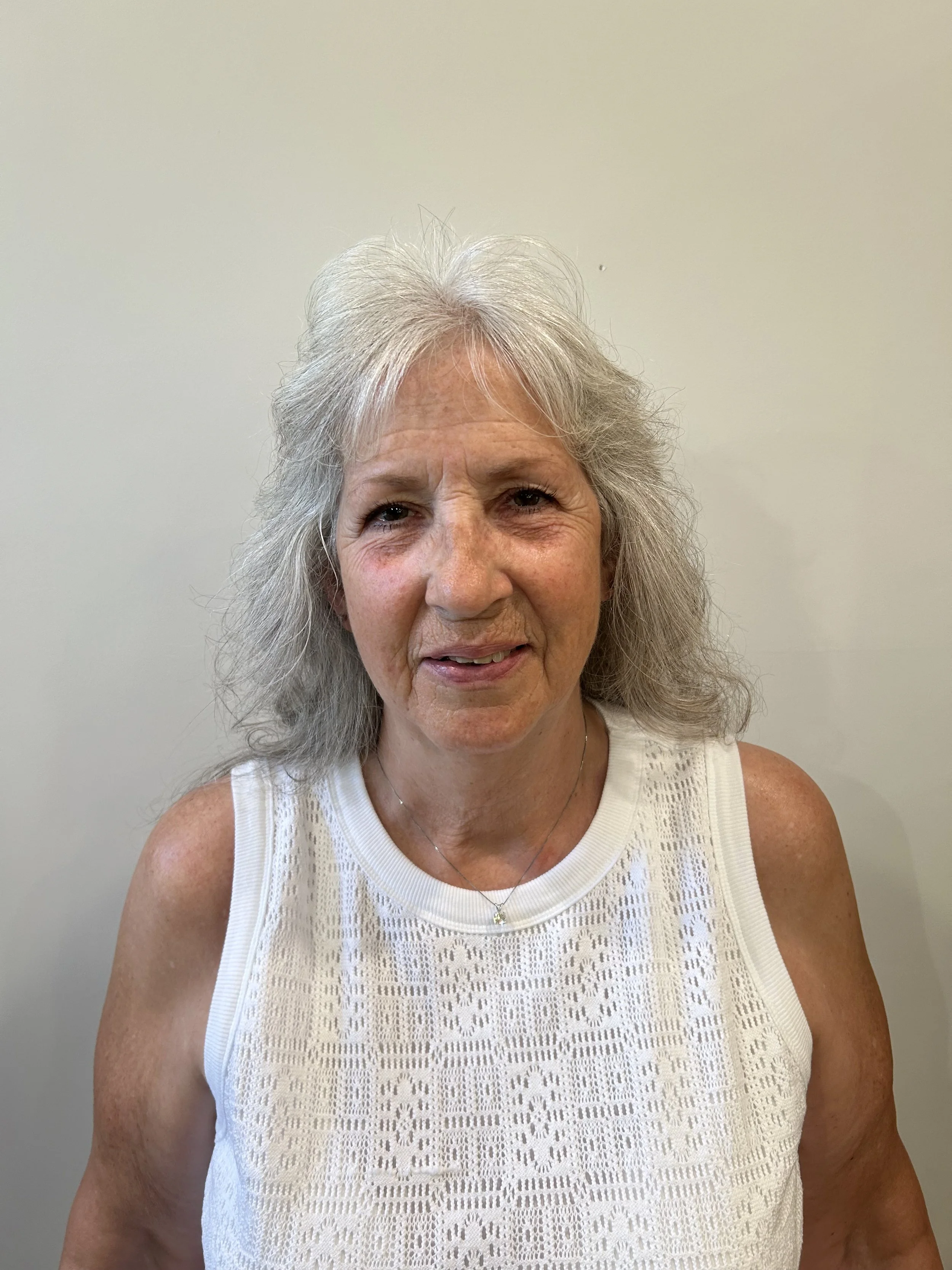 A middle-aged woman with gray, wavy hair smiling at the camera, wearing a white sleeveless textured top and a delicate necklace, standing against a plain, light-colored wall.