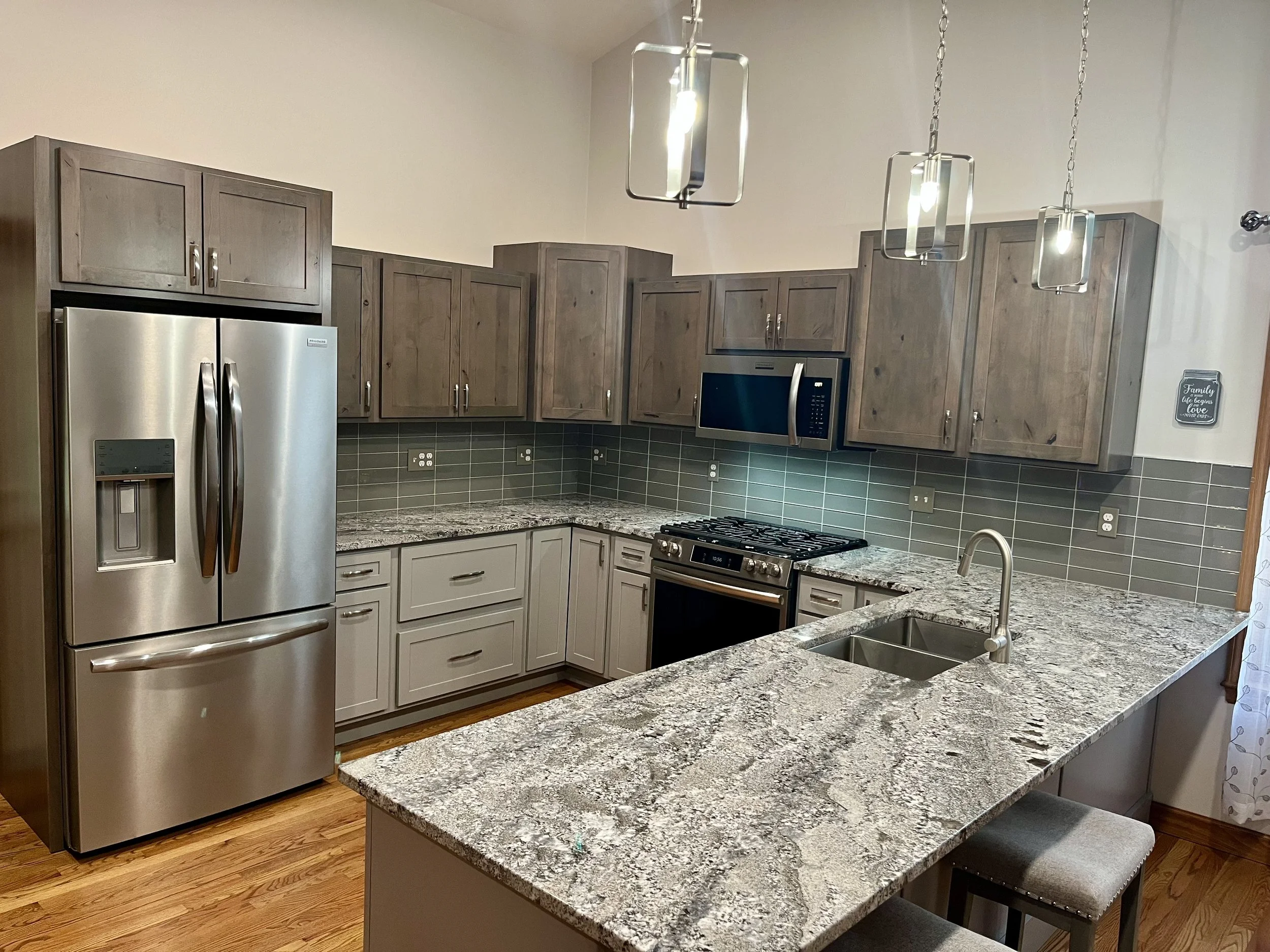 Kitchen with gray wooden cabinets, stainless steel refrigerator, granite countertops, built-in microwave, gas stove, undermount sink, tiled backsplash, light fixtures hanging from the ceiling, wooden flooring, and a small decorative sign on the wall.