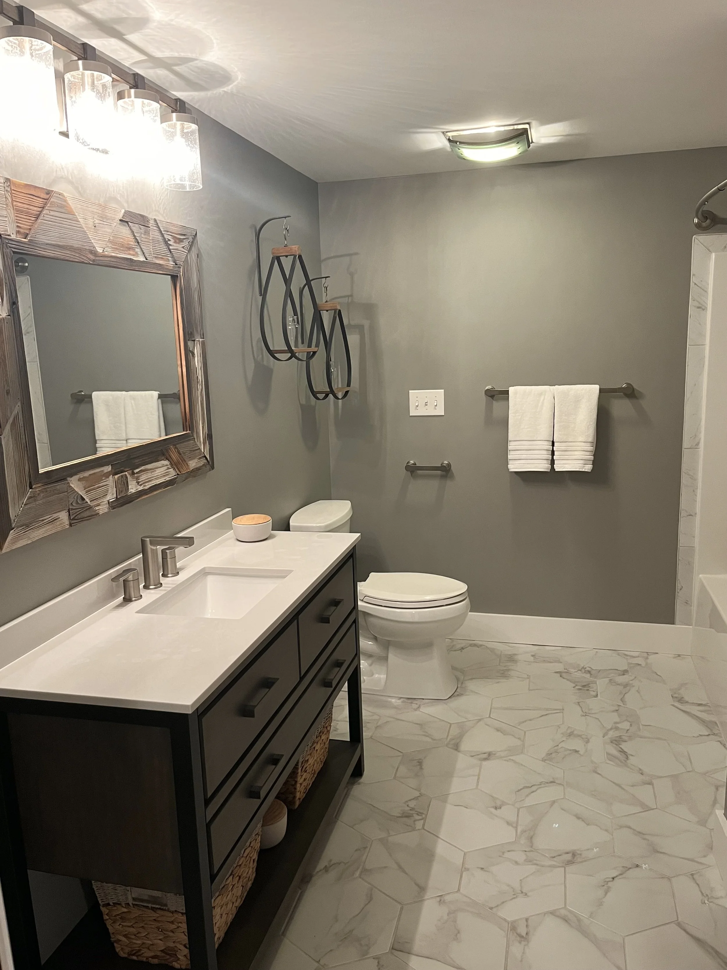 Modern bathroom with gray walls, white marble floor, black vanity with wicker baskets, mirror with wooden frame, white towel on silver towel bar, decorative wall black metal shelves, white toilet, ceiling light, and a shower with marble walls.