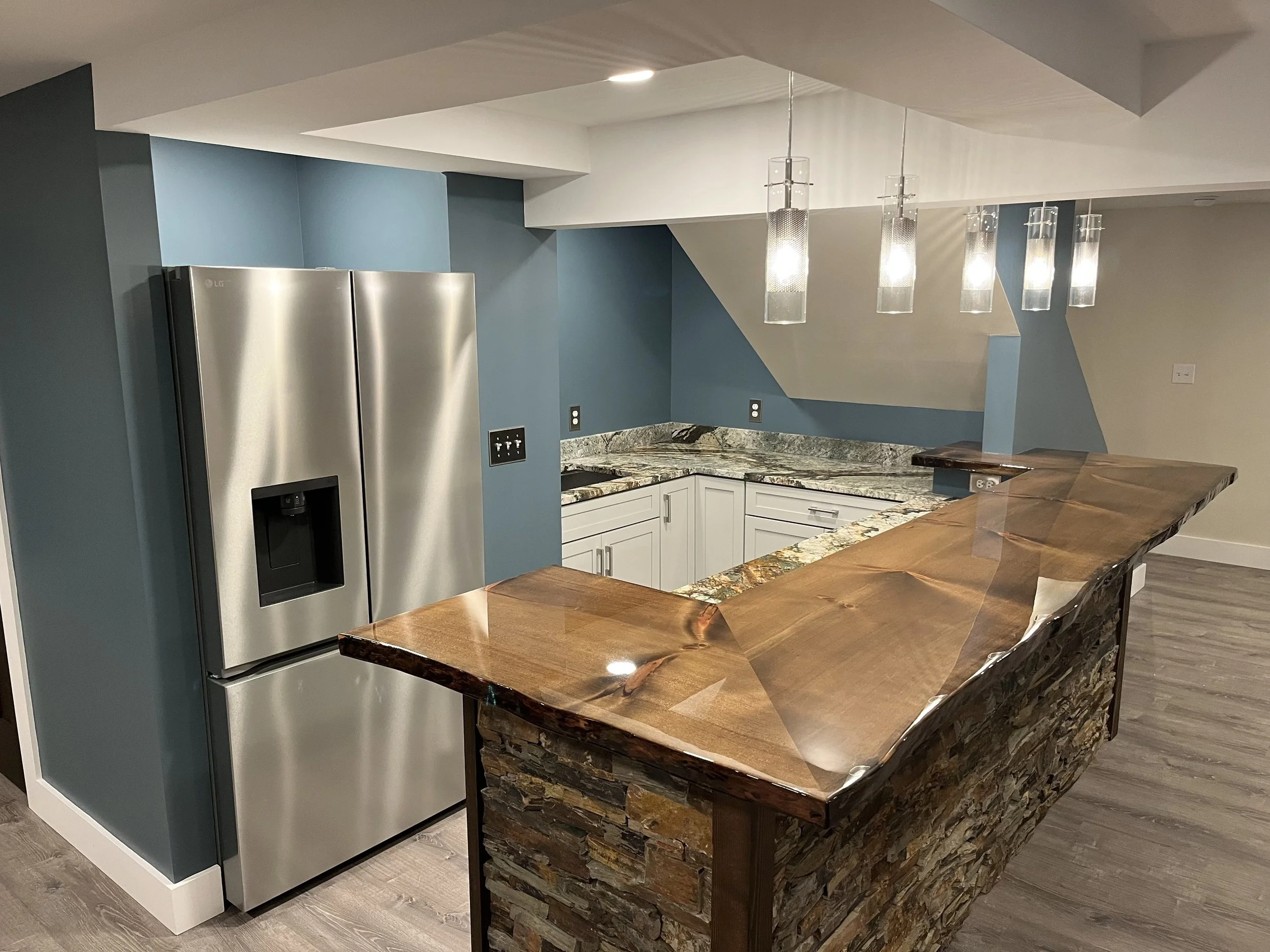 Modern kitchen with blue and white walls, stainless steel refrigerator, granite countertops, and a wooden kitchen island with stone accents, hanging pendant lights, and electrical outlets.