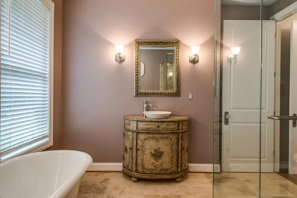 Bathroom with a vintage vanity, oval mirror, wall sconces, a white vessel sink, a clawfoot tub, a window with blinds, and a glass shower enclosure.