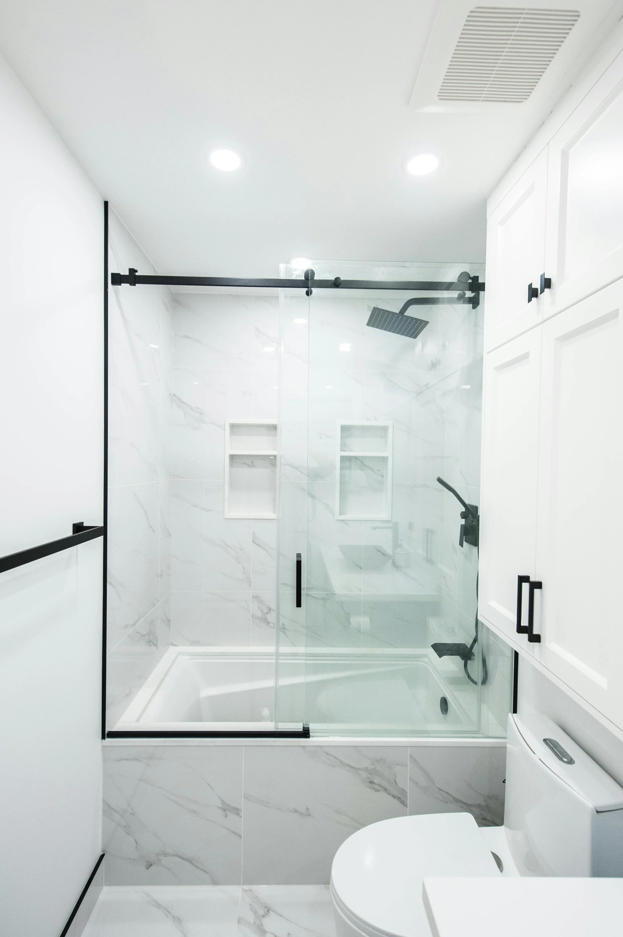 Modern bathroom with white marble walls, a glass shower enclosure with black hardware, ceiling lights, a wall-mounted showerhead, a white cabinet, and a white toilet.